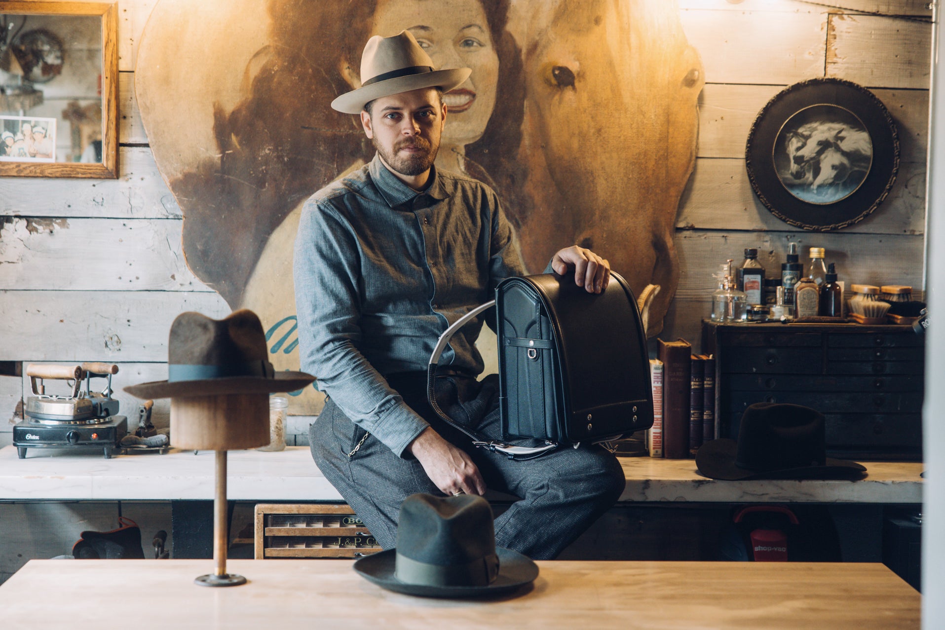 John Fish wearing a fedora and a blue shirt, holding a black Randoseru backpack, sits on a counter in a rustic room adorned with a large painting of a woman and horses, with other hats displayed in the foreground.