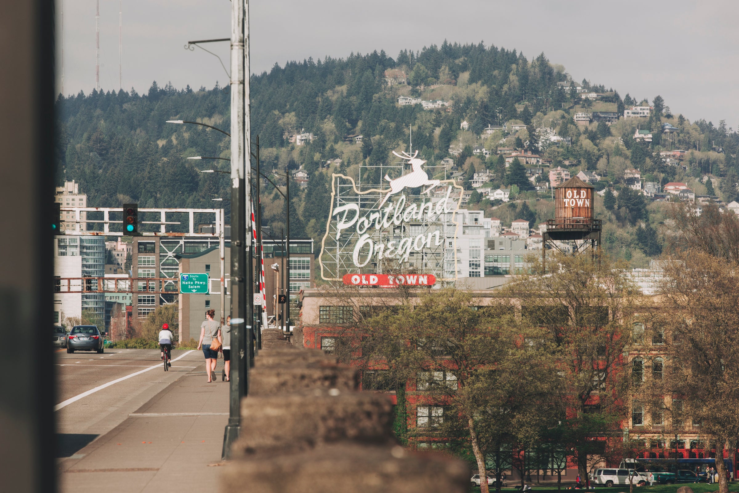 A cityscape photo of Portland, Oregon, showing the iconic "Old Town" sign with a white stag, with a street and a tree-covered hill in the background.