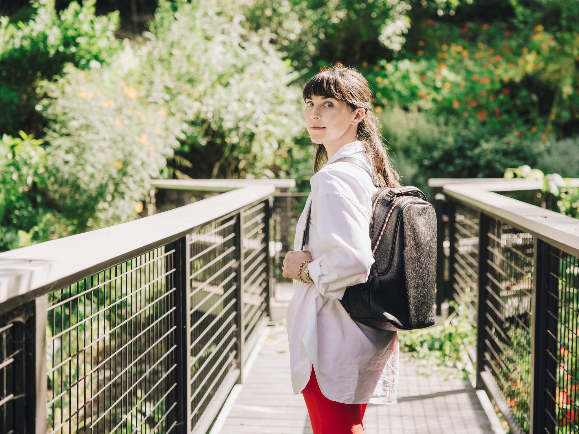 Tsuchiya Kaban's Ambassador's Julia Sherman with dark hair and bangs looks over her shoulder, wearing a white collared shirt, red pants, and a Tsuchiya Kaban's Clarte Backpack in Black, standing on a wooden walkway with metal railings surrounded by green trees and foliage in the background.
