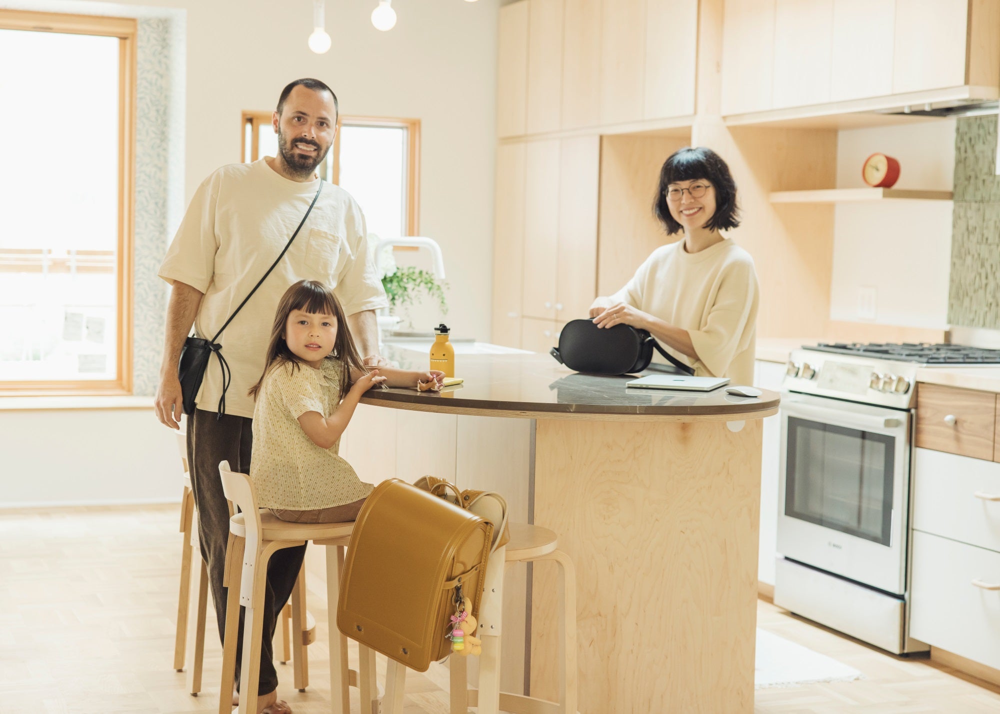 A smiling family of three – Tsuchiya Kaban’s ambassador Airi Isoda, Ryan Upton and their daughter Kenzo– are in a modern kitchen with light wood cabinets. Ryan and Kenzo stand near a counter with a Tsuchiya Kaban’s Randoseru Lite in yellow hanging from a stool, while Airi sits at the counter with a Tsuchiya Kaban’s Black Nume Oval Shoulder in Black.