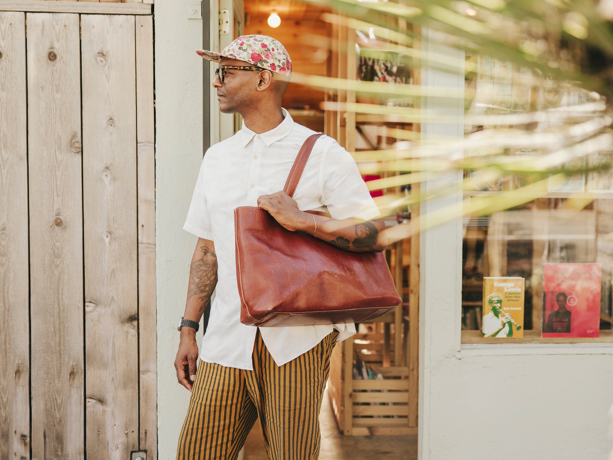 Dário Solari stands outside a building, looking to his left, wearing a white short-sleeved collared shirt, striped pants, a floral cap, and glasses, while carrying a Tsuchiya Kaban's Diario Large Tote in Brown on his right shoulder. 