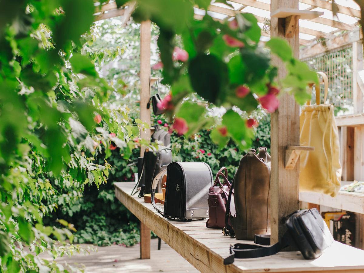 Various Tsuchiya Kaban’s leather handbags and backpacks are arranged on a wooden table, under a canopy of lush green leaves and some small pink flowers, suggesting an outdoor market or garden setting.