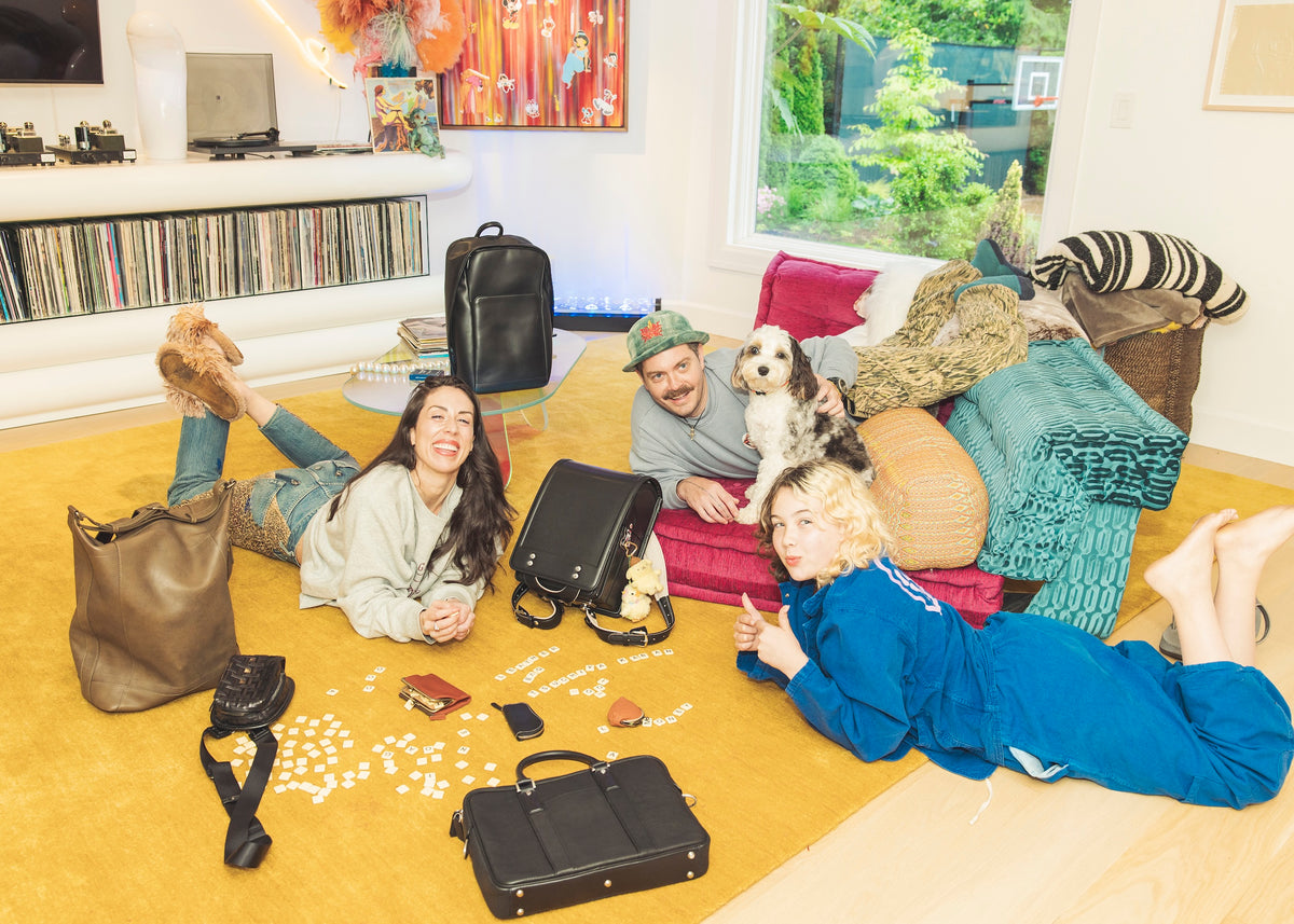 A cheerful group of three people and a dog are lying on a yellow rug in a bright living room. A woman with long dark hair is on the left, smiling at the camera. In the center, a man with a mustache and a hat is lying next to a small dog, also smiling. A woman with short blonde hair and a blue jumpsuit is on the right, giving a thumbs-up. Several leather bags, wallets, and what looks like a board game are scattered around them on the rug.