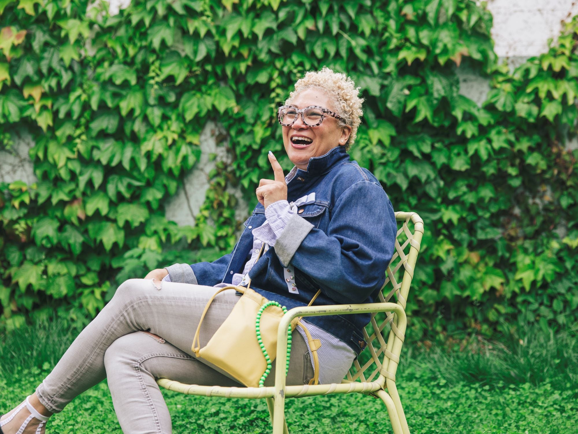 Kimiko Matsuda with short, curly blonde hair and glasses sits in a light green chair, smiling and pointing up, with a Tsuchiya Kaban’s Ruffle Drawstring Pouch in Yellow beside her and green ivy in the background.