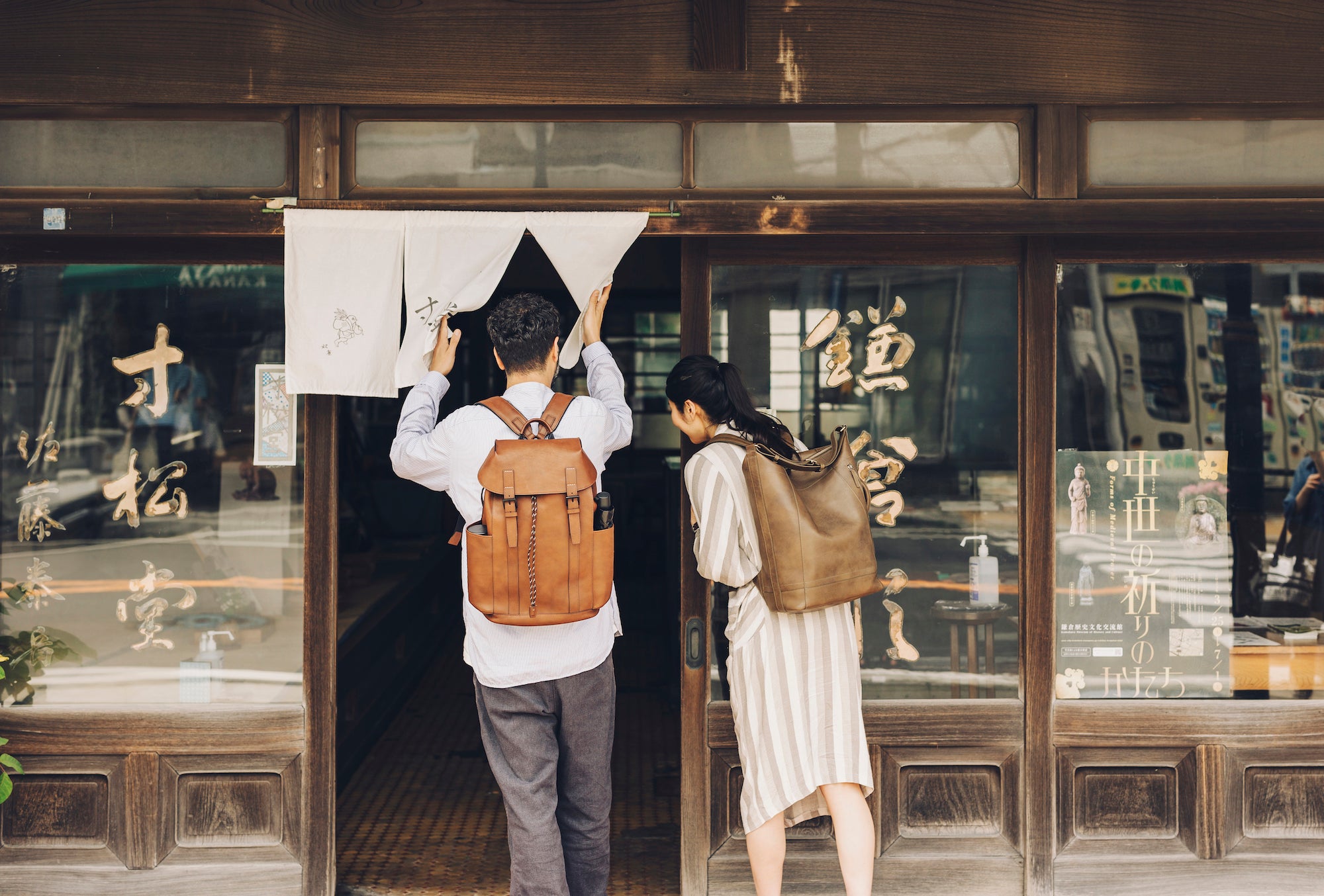 Omar with a Tsuchiya Kaban's Large Twin Belt Backpack adjusts a white noren curtain in the doorway of a traditional Japanese building, while Kayo with a Tsuchiya Kaban's  Roadster 3way Backpack ooks into the shop next to him.