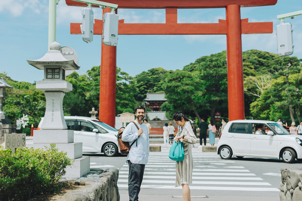 Omar with a Tsuchiya Kaban's  Large Twin Belt Backpack smiles at the camera while standing on a crosswalk next to Kayo looking into her Tsuchiya Kaban's Clarte Gathered Shoulder with a large red torii gate and lush green trees in the background.