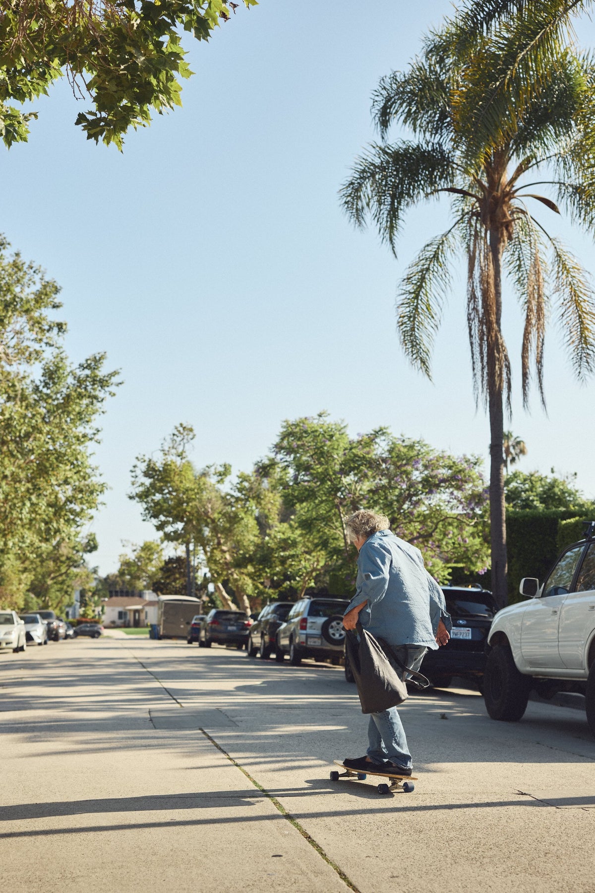 Steve rides a longboard skateboard down a sunny residential street lined with parked cars, palm trees, and green foliage under a clear blue sky, with his Tsuchiya Kaban leather Rolltop Backpack.
