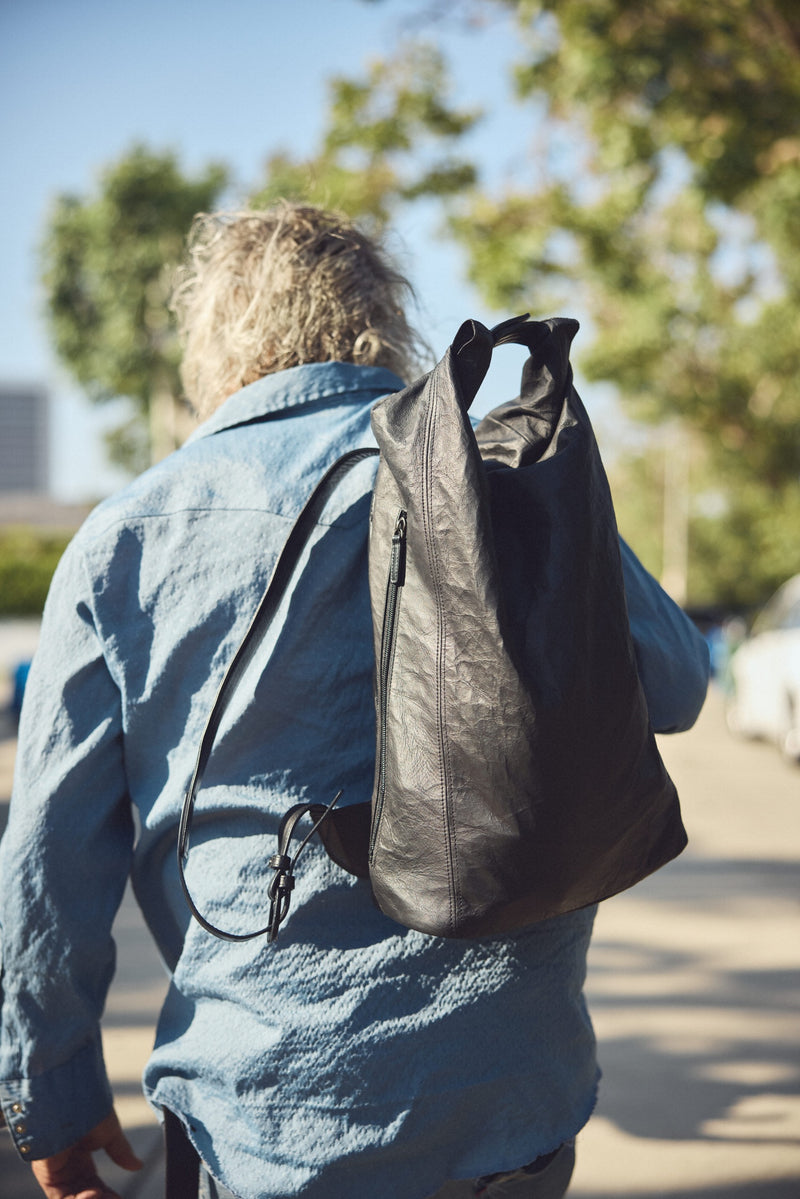 Steve Olson seen from behind wearing a light blue denim shirt and carrying a Tsuchiya Kaban's black leather Rolltop backpack outdoors on a sunny day.
