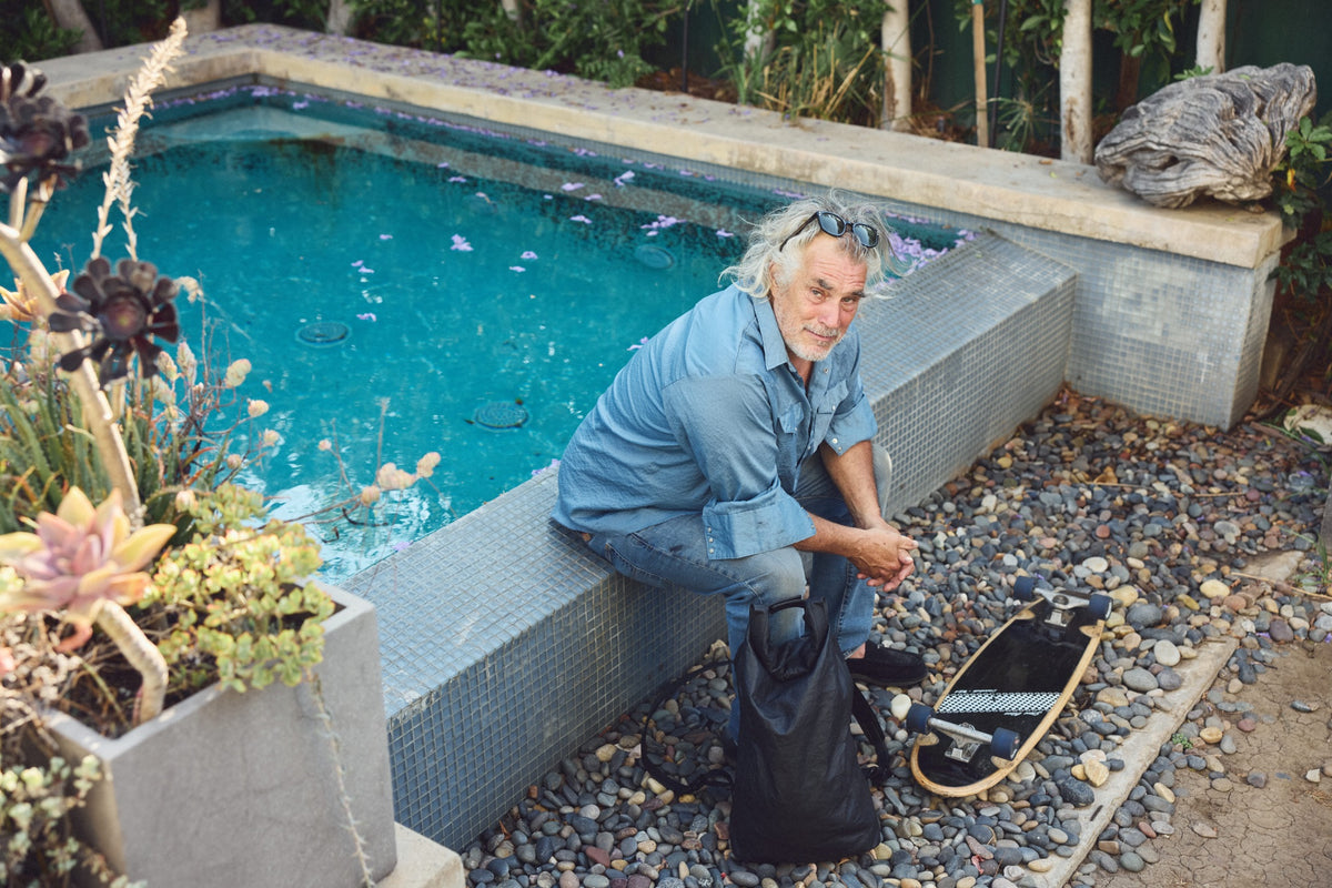 Steve Olson sits on a tiled poolside ledge wearing a light blue denim shirt and jeans, with a black Tsuchiya Kaban Rolltop backpack on the ground beside him and a skateboard nearby. The setting features a swimming pool, gravel, plants, and a stone wall in the background.