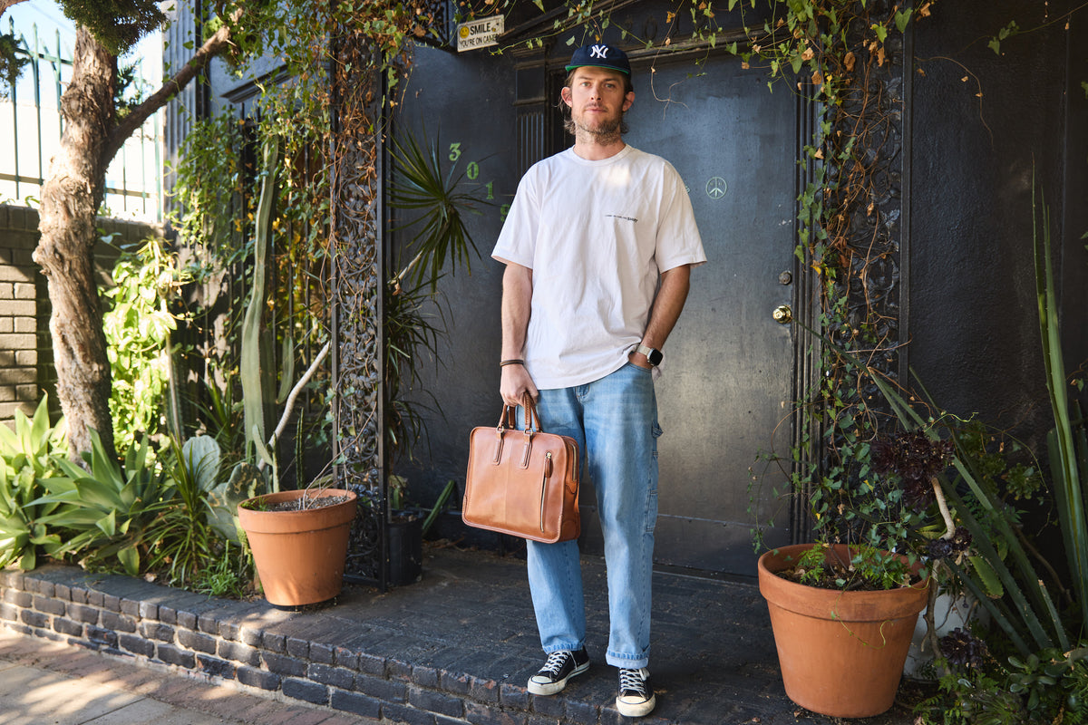Tsuchiya Kaban’s Ambassador, Ariel Stark, with a light beard and a dark baseball cap is standing on a concrete step in front of a dark, vine-covered building. He is wearing a loose-fitting white t-shirt, light-wash jeans, and dark sneakers with white laces. In his right hand, he holds a Tsuchiya Kaban’s Urbano Urban Briefcase in Brown. To his left and right are large terracotta pots with green plants. 