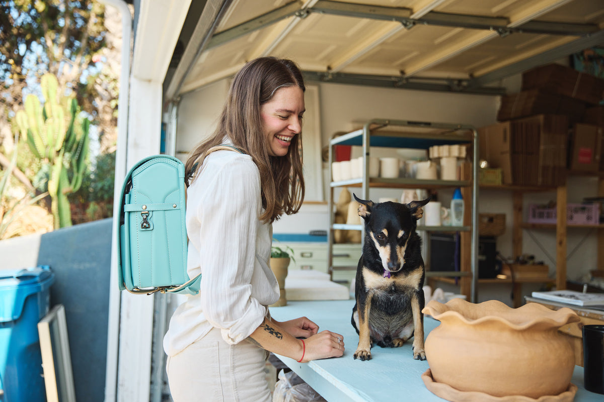Haley Bradley, a smiling woman with long brown hair, wears a light-colored top and pants, and carries a light blue Tsuchiya Kaban’s Randoseru Cowhide backpack. She is leaning over a table, interacting with a small black and tan dog sitting on it, with pottery supplies and a garage door in the background.