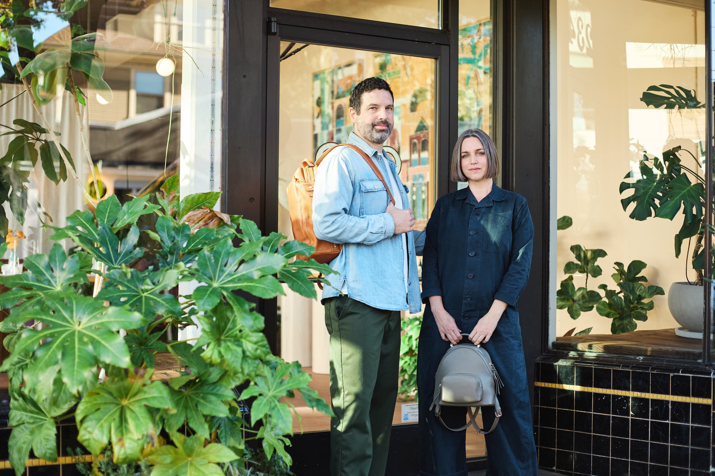 Tsuchiya Kaban’s ambassador Heather and Jonathan Sielaff stand outside a storefront adorned with plants. Jonathan has a beard and dark hair with some grey, wearing a light blue denim shirt, dark green pants, and a Tsuchiya Kaban Tone Nume Medium Backpack in Medium Brown. Heather has short grey hair and wears a dark blue jumpsuit. She is holding a Tsuchiya Kaban Clarte Small Backpack in Ash Gray.