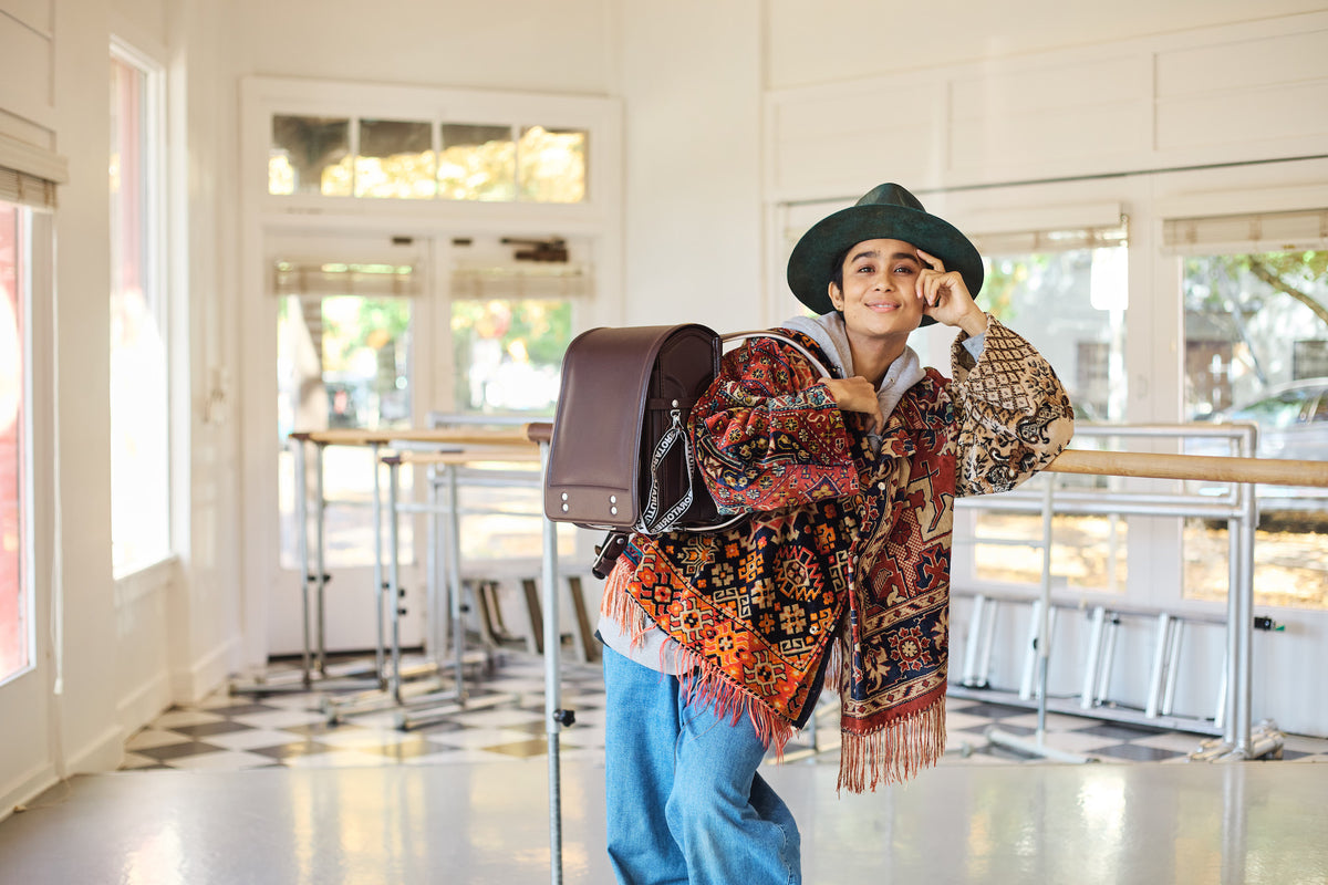 Tsuchiya Kaban’s ambassador UNO wearing a colorful patterned jacket and green hat leans on a railing indoors, smiling, with a Tsuchiya Kaban’s Brown Randoseru Cowhide over one shoulder.