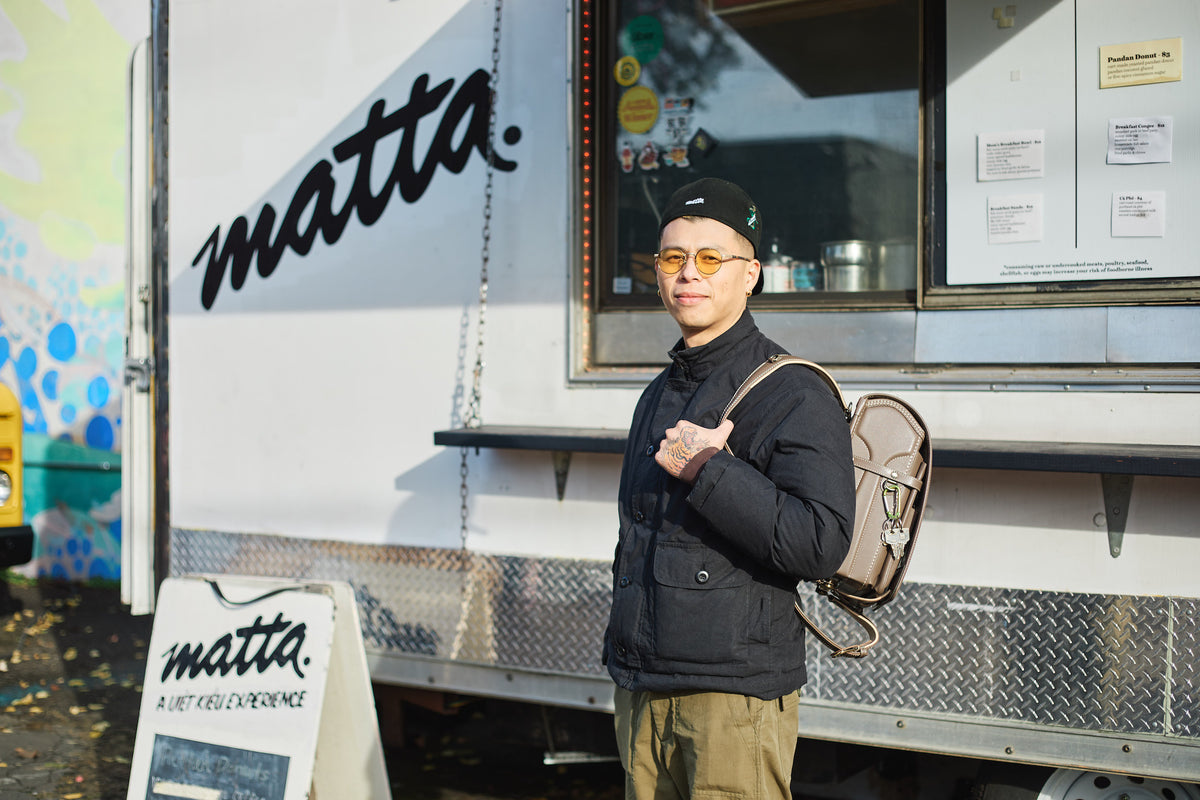 Tsuchiya Kaban’s ambassador Richard Le stands in front of a “matta” food truck, wearing a black jacket and cap, carrying a Tsuchiya Kaban's Randoseru over one shoulder.
