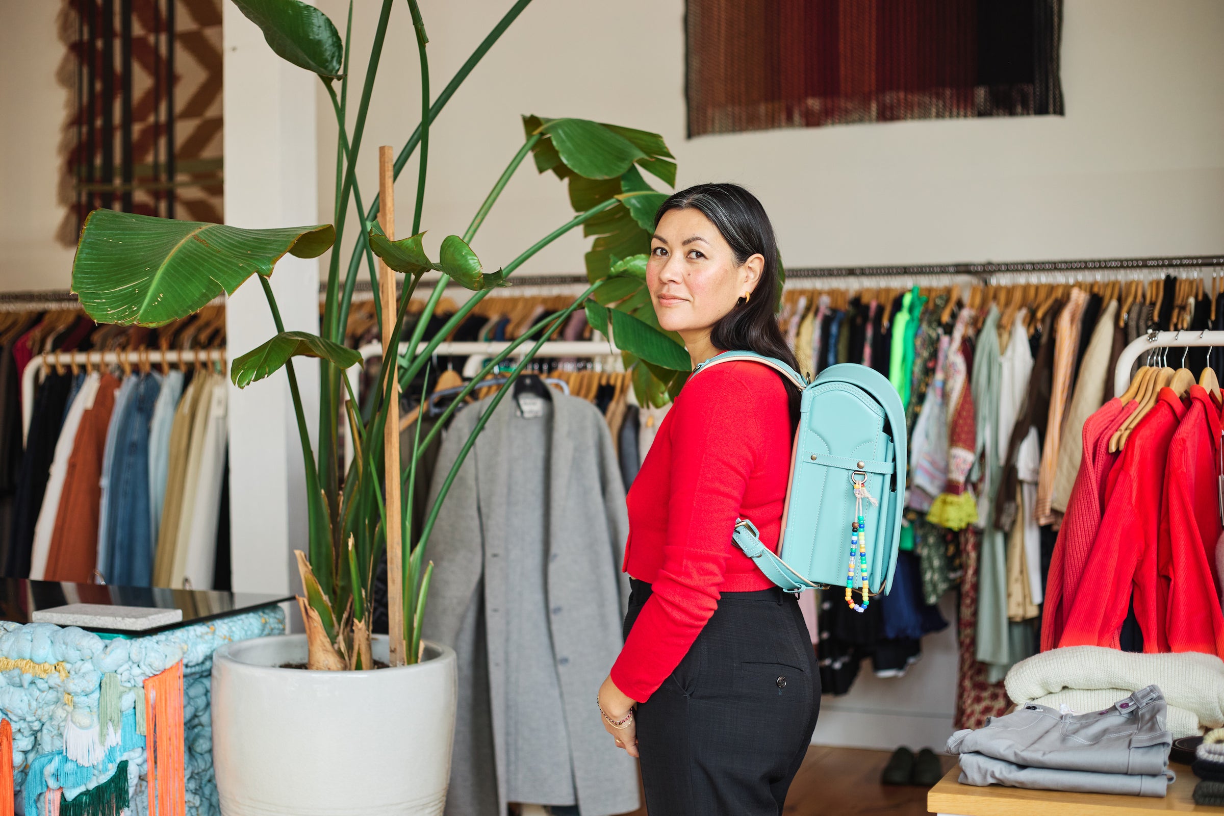 Pamela Baker-Miller, founder and owner of Frances May, standing in a stylish clothing boutique wearing a red top and carrying a light blue Randoseru.