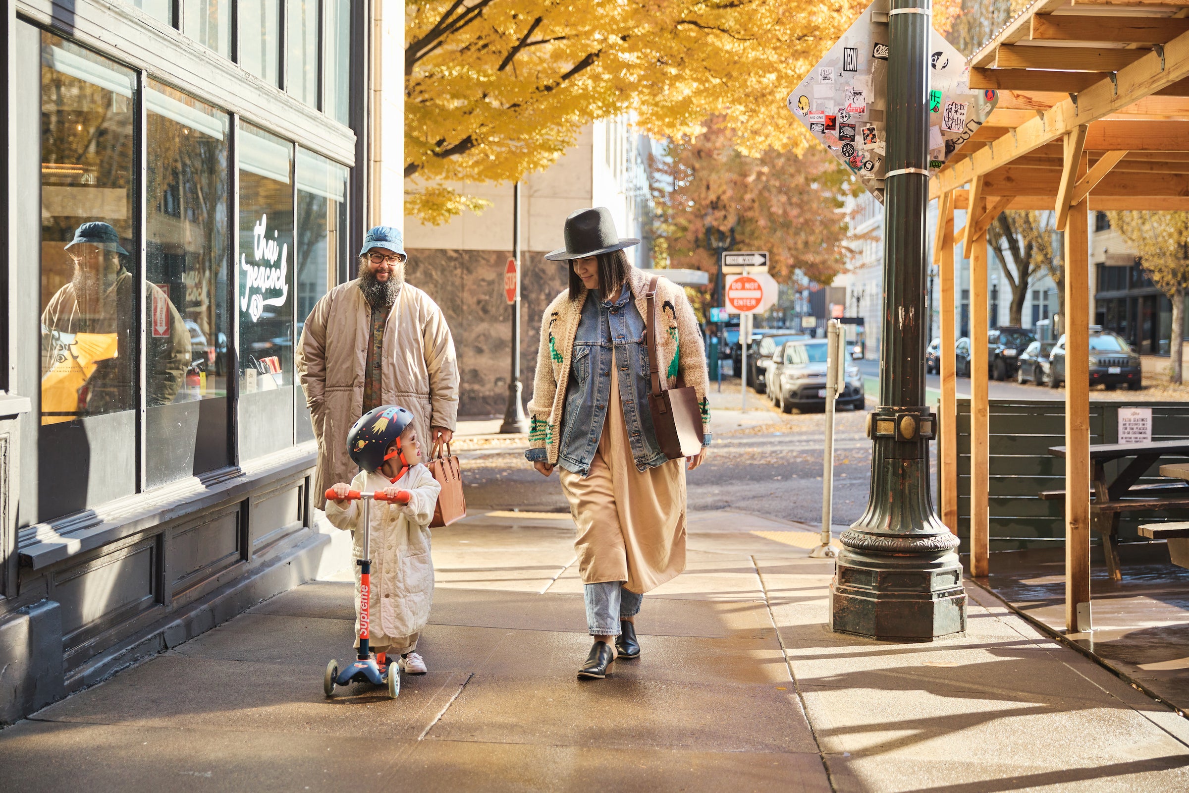 A family of three walks down a city sidewalk on a sunny day. Tsuchiya Kaban’s ambassador  Ashley Thornton in a black hat, denim jacket, and long beige dress walks in the foreground, carrying a Tsuchiya Kaban’s Original Nume Vase Shoulder and a book. Beside them, a young child in a light-colored jumpsuit and helmet rides a scooter. Behind them, Colton Tong of Tsuchiya Kaban’s ambassador with a beard and a light brown jacket smiles. The sidewalk is lined with buildings and trees with yellow autumn leaves.