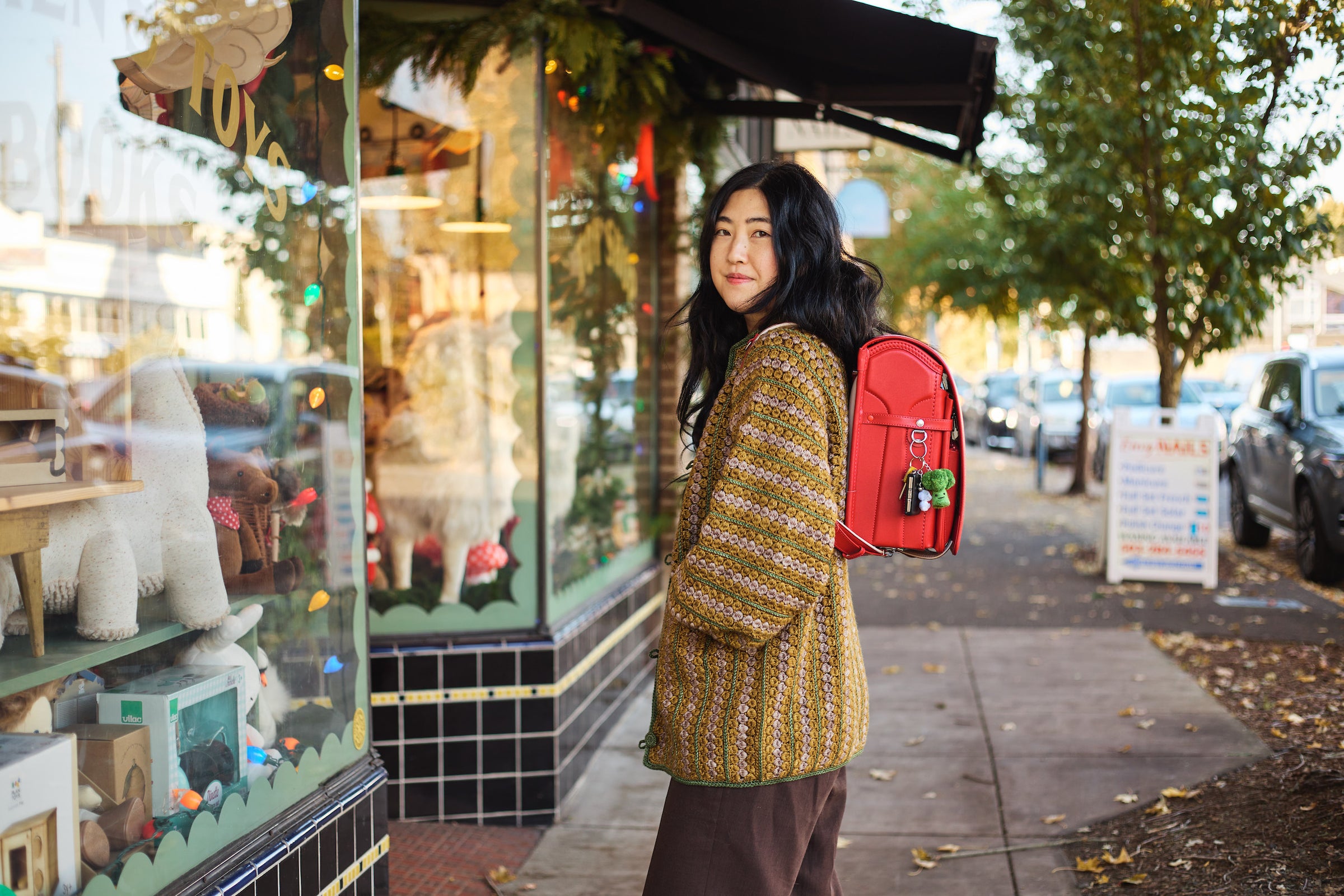 Tsuchiya Kaban’s ambassador Caroline Kim Rodrigues, owner and founder of Shop Merci Milo, standing outside a storefront and facing the showcase with her body but looking back toward the camera with a slight smile while wearing a Tsuchiya Kaban’s Red Randoseru Cowhide and a cozy knit sweater.