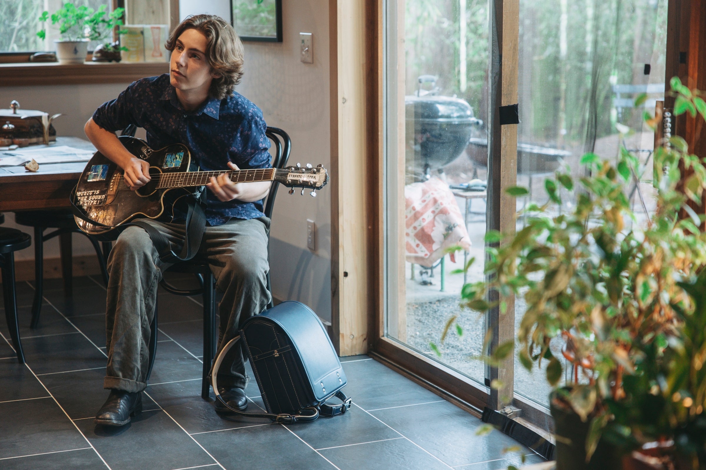 A young man sits on a chair in a room with a large window and plays an acoustic guitar. A Tsuchiya Kaban's Randoseru in Black rests on the floor next to him.