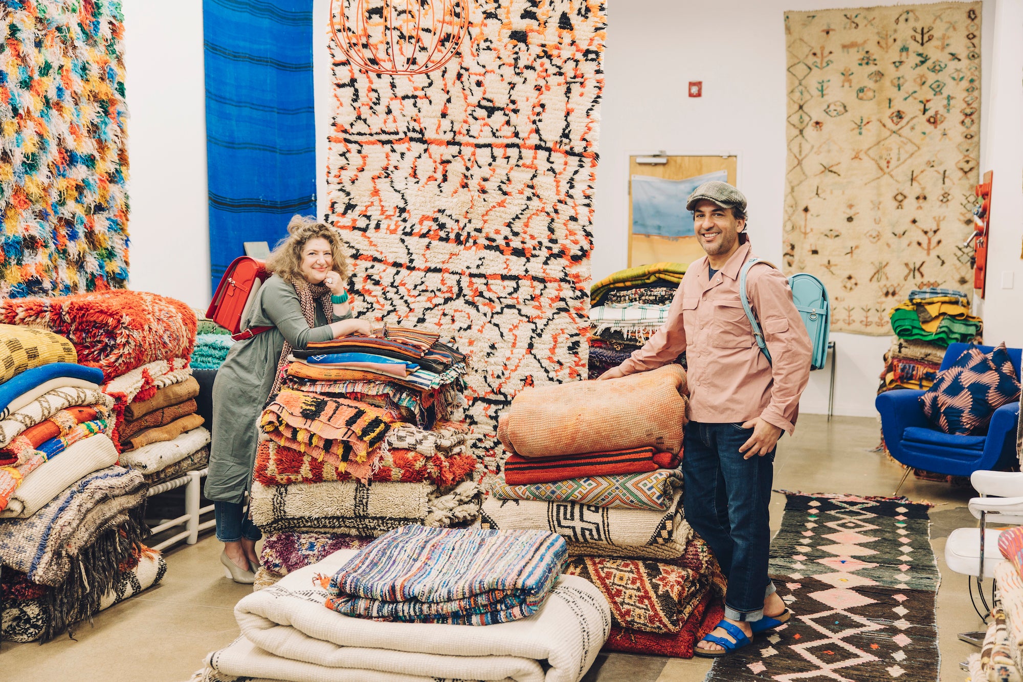 Katen with a Tsuchiya Kaban's Red Randoseru and  Latif in a cap with a Tsuchiya Kaban's Blue Randoseru stand among large stacks of colorful rugs and textiles in a showroom.