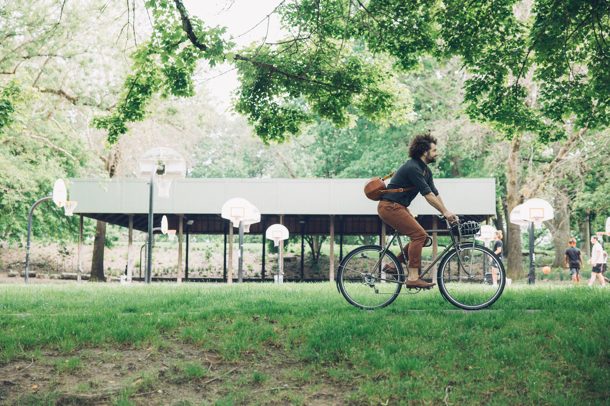 Adam Porterfield rides a bicycle across a grassy area, wearing a dark shirt, brown pants, and a round brown bag slung over his shoulder. In the background, there's an outdoor basketball court with multiple hoops and a few people. The scene is set under a canopy of green trees.