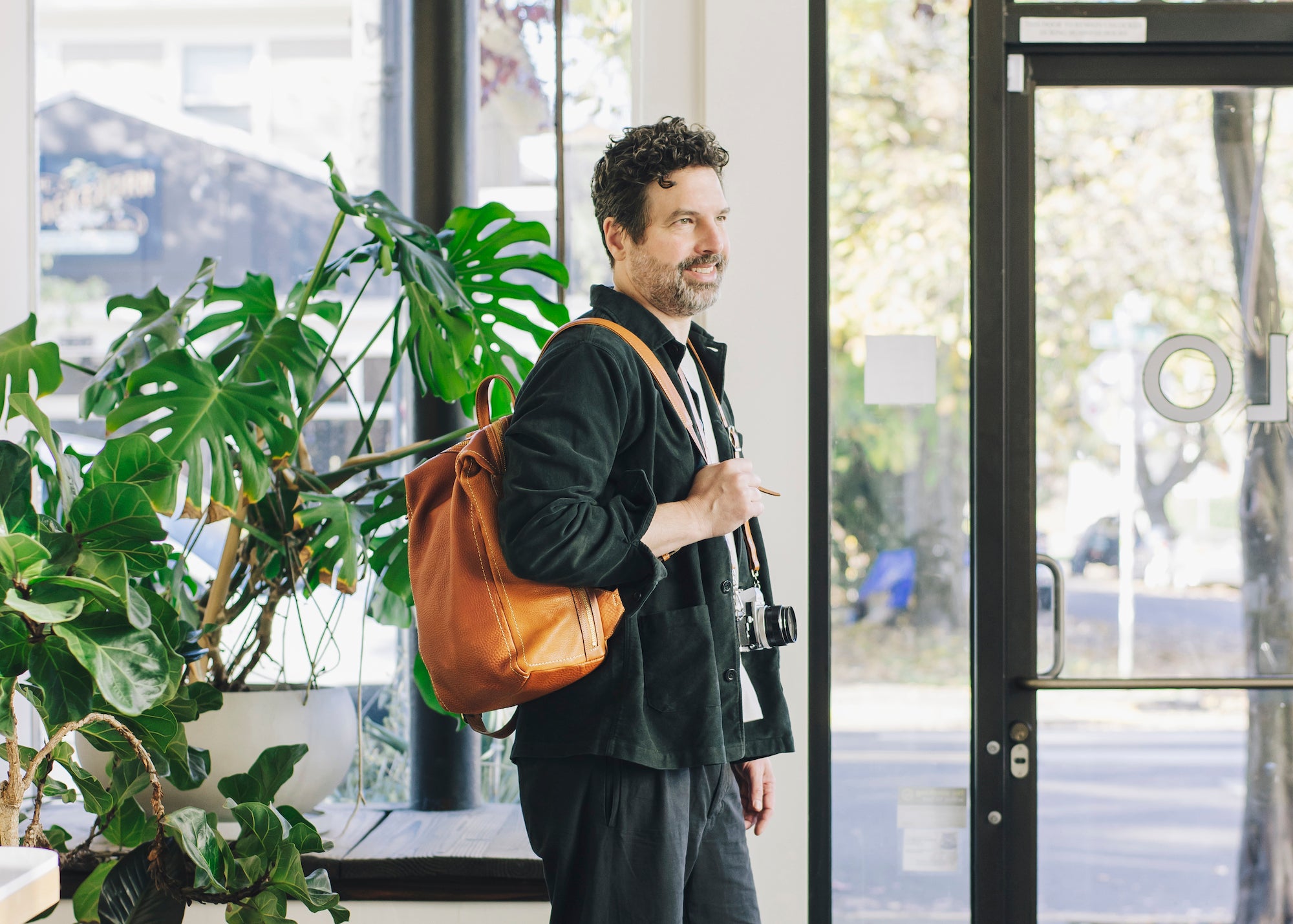 Jonathan Sielaff, a man with a beard, wears a dark shirt and carries a Tsuchiya Kaban’s Tone Nume Medium Backpack and a camera, standing indoors next to a large plant and looking out a glass door.