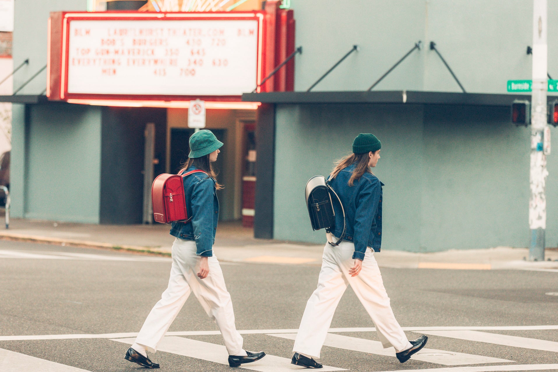 Two individuals, one with a red backpack and a green hat, and the other with a black backpack and a green hat, walk across a street in front of a theater with a marquee sign.