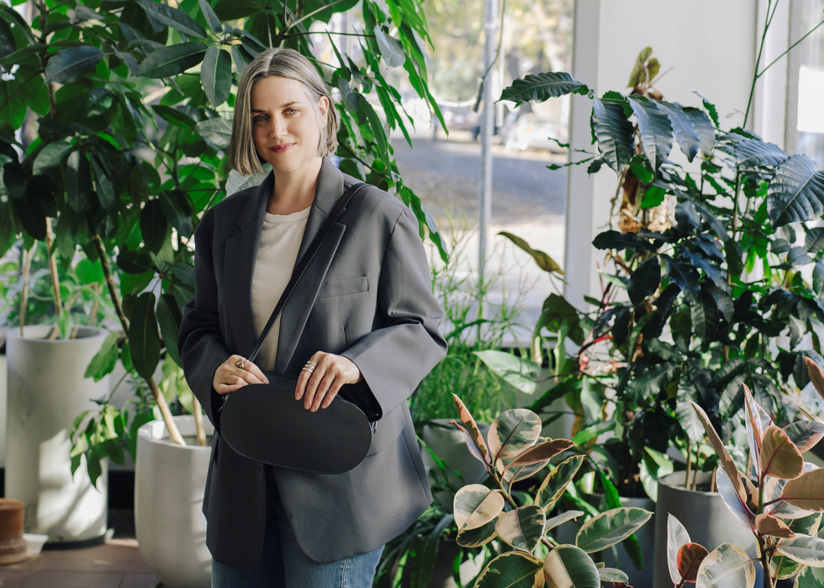 Heather Sielaff with short, light brown hair styled in a bob stands in a room filled with potted plants. She is wearing a grey oversized blazer over a light-colored t-shirt and what appears to be blue jeans. She holds a a Tsuchiya Kaban’s Black Nume Oval Shoulder in Black with a long strap in front of her.