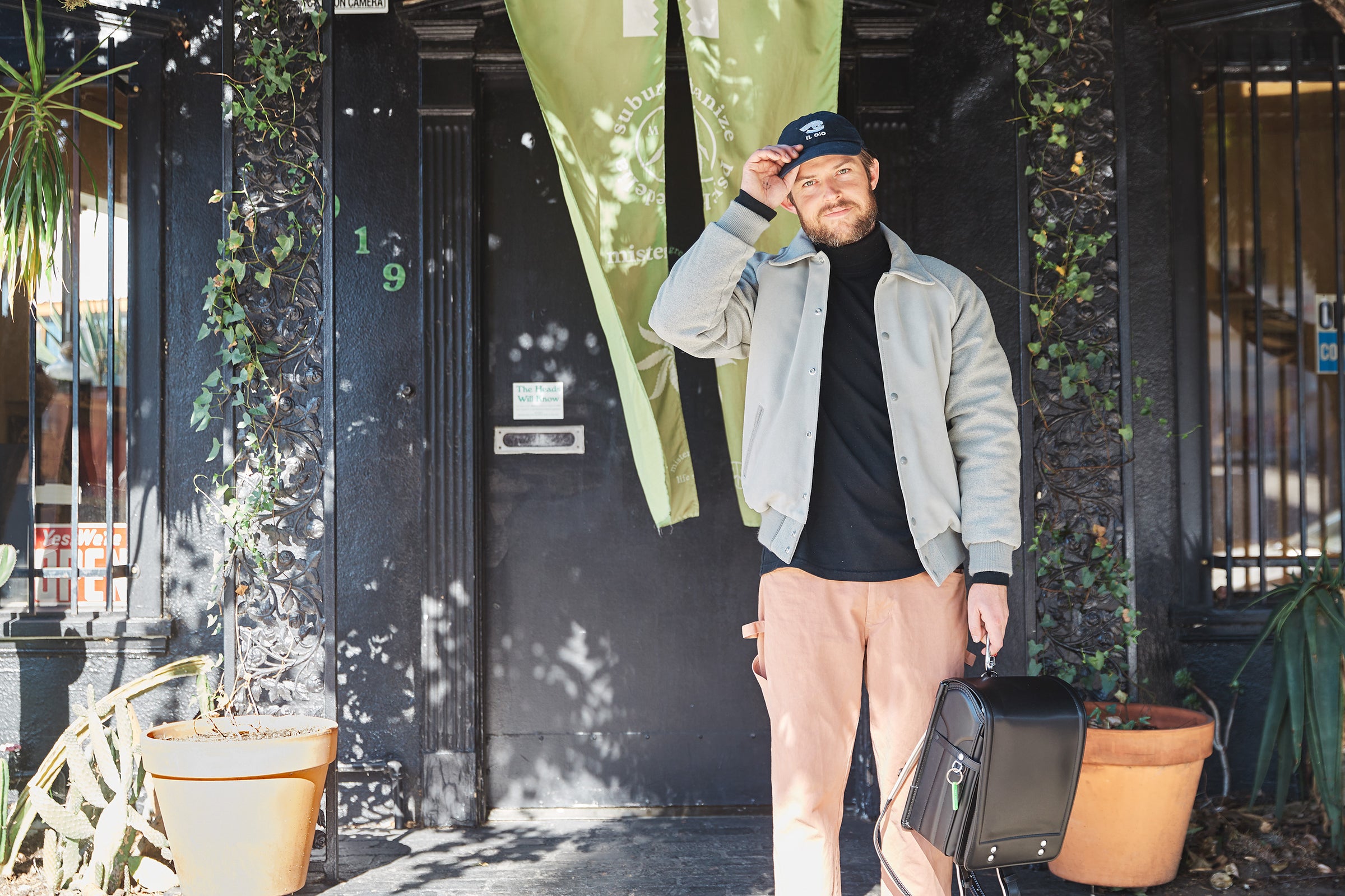 Ariel Stark-Benz, founder and owner of Mister Green, standing in front of a black storefront with green banners, wearing a light jacket and cap, and holding a Black Randoseru Cowhide.