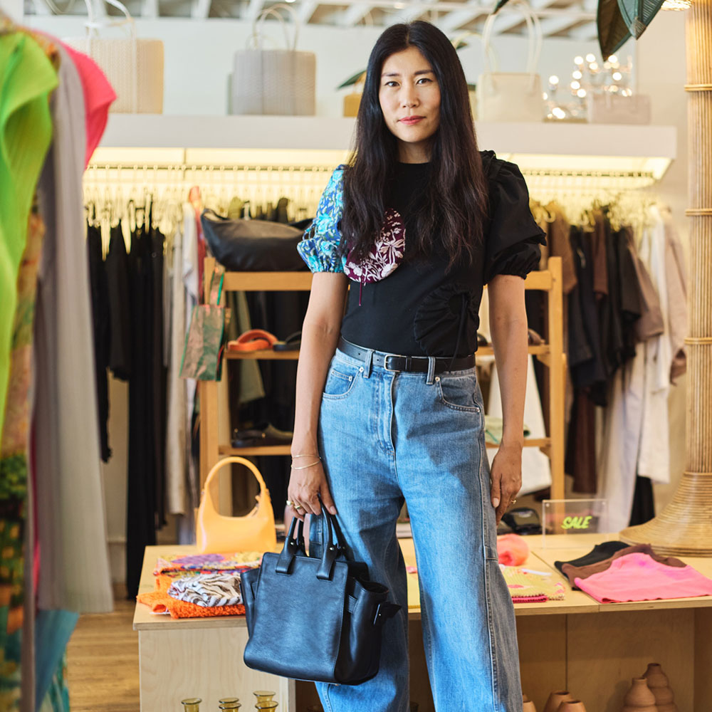 Tsuchiya Kaban’s ambassador Bo Carney standing in a clothing store wearing a black t-shirt with colorful puffed sleeves and high-waisted blue jeans, holding a Tsuchiya Kaban’s Editor Bloom Tote in Black. Shelves of shoes and racks of clothes are visible in the background.