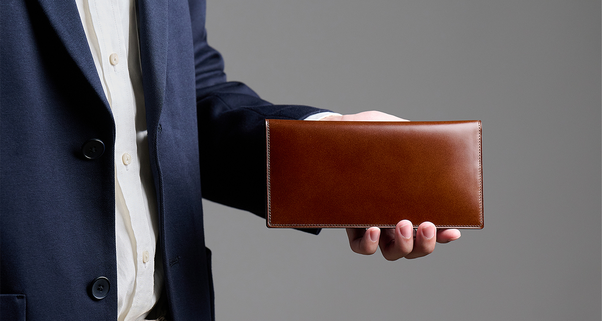 A person wearing a dark suit jacket and a white shirt holds a Tsuchiya Kaban's Cordovan Long Wallet.