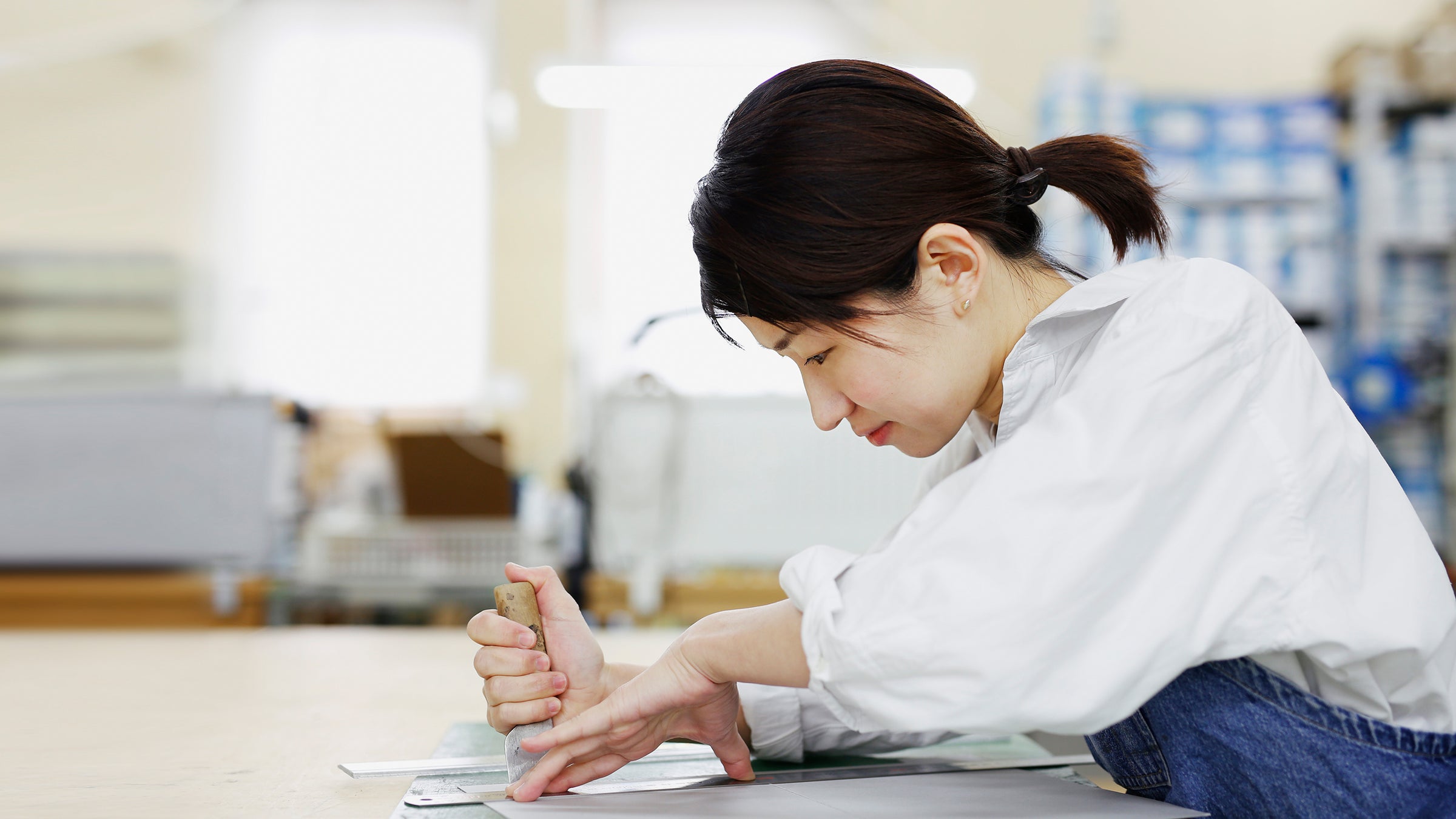 A side profile shot of a woman in a white shirt and jeans, with her hair in a ponytail, carefully cutting a piece of material on a table in a workshop.