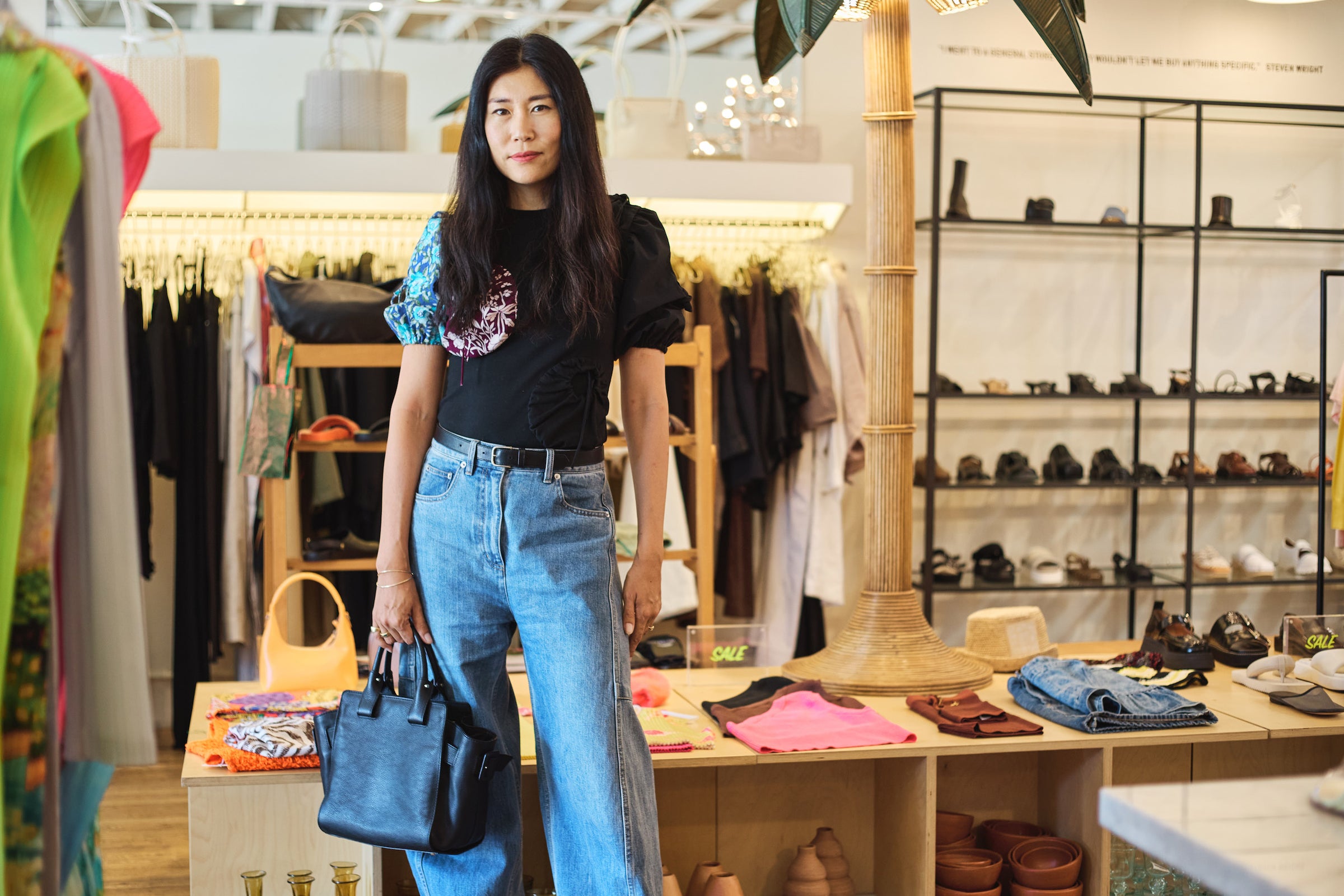 Tsuchiya Kaban’s ambassador Bo Carney standing in a clothing store wearing a black t-shirt with colorful puffed sleeves and high-waisted blue jeans, holding a Tsuchiya Kaban’s Editor Bloom Tote in Black. Shelves of shoes and racks of clothes are visible in the background.