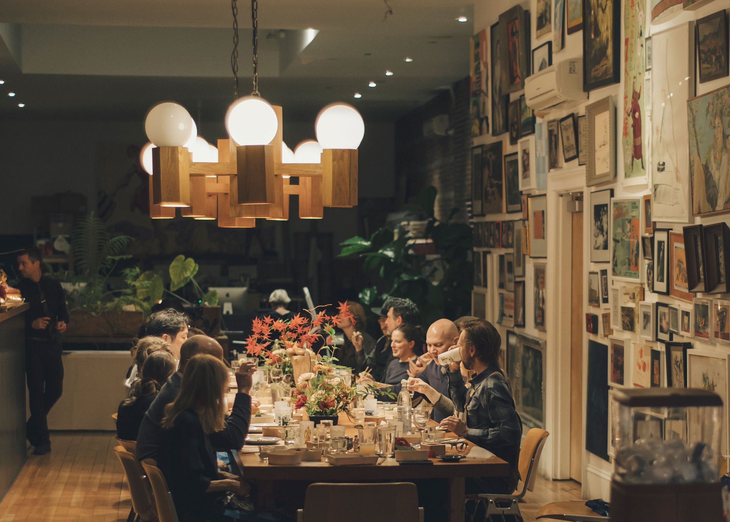 A group of people are gathered around a long dining table in a warmly lit restaurant or private dining space. A striking wooden chandelier with globe lights hangs above the table, which is adorned with flower arrangements. 