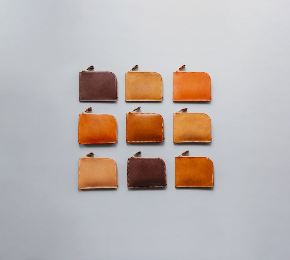 Tsuchiya Kaban's  perfectly aligned grid of nine small, rectangular leather  wallets is displayed . The pouches come in a gradient of brown and tan hues, ranging from darker browns to lighter, more golden tones, and each features a small zipper closure with a leather pull tab.