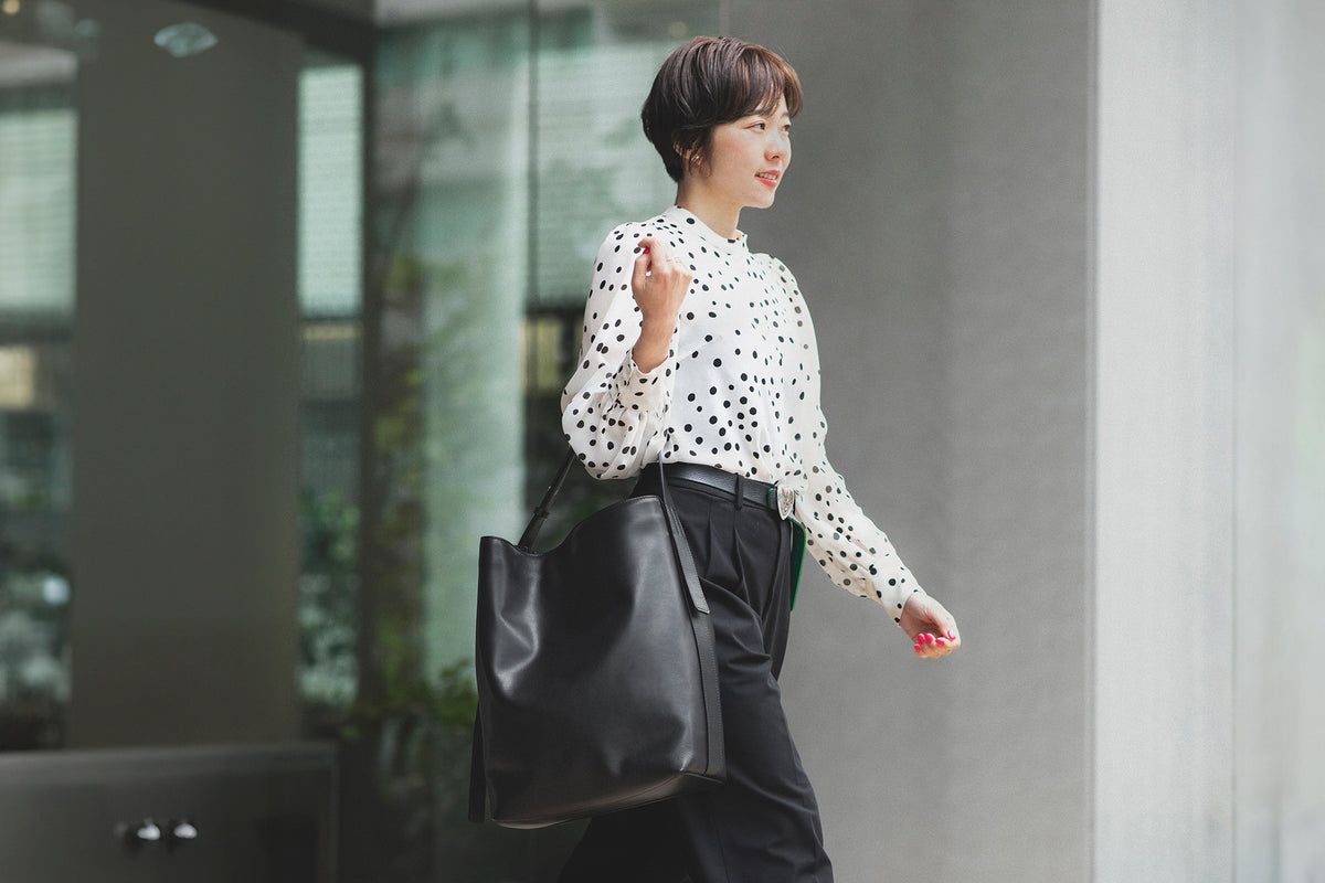 A woman walking outdoors carrying a large black Atlow leather bucket tote bag from Tsuchiya Kaban. She is wearing a white long-sleeve blouse with black polka dots and black trousers.