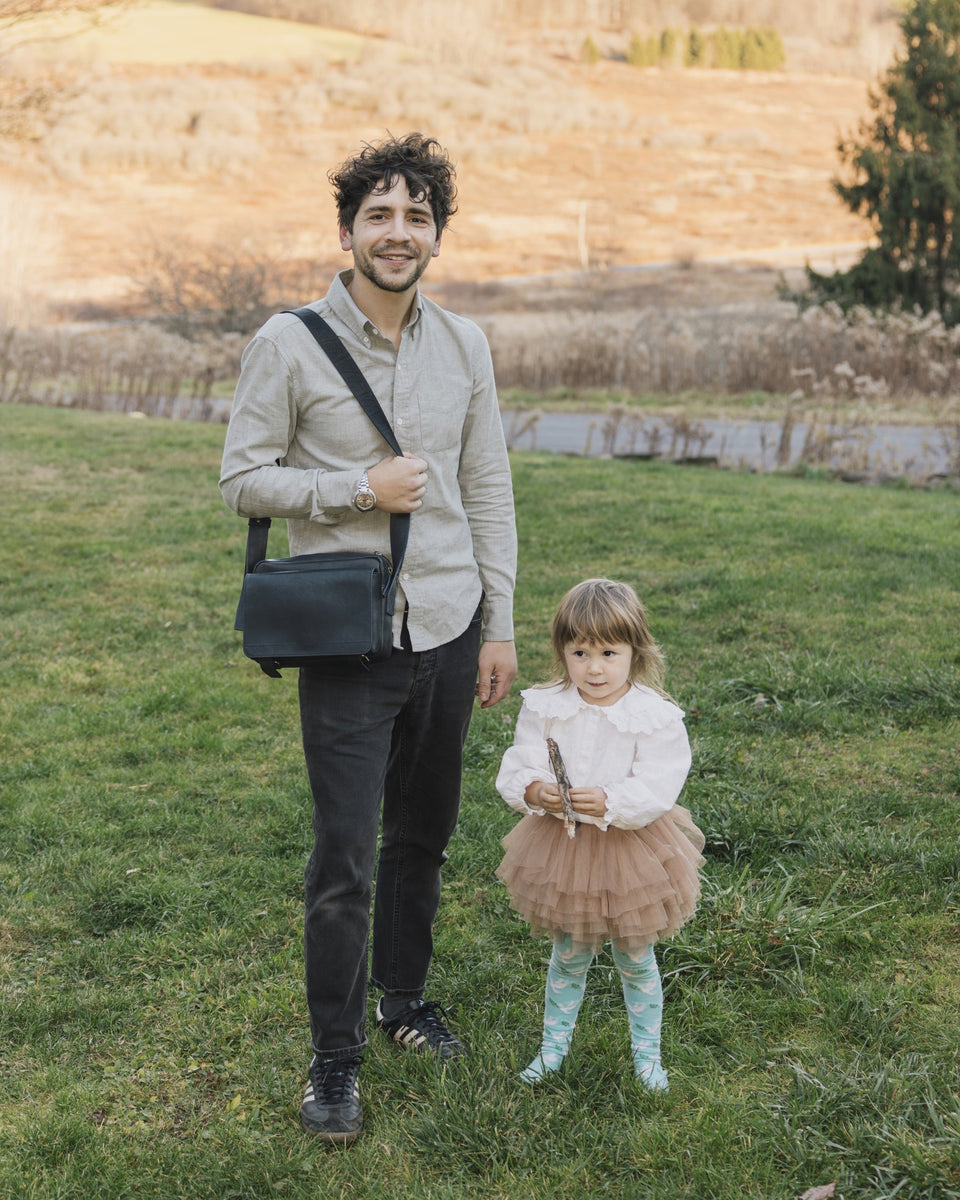 Father with a dark navy Roadster leather bag standing next to a young girl, his daughter in a dress on the grass