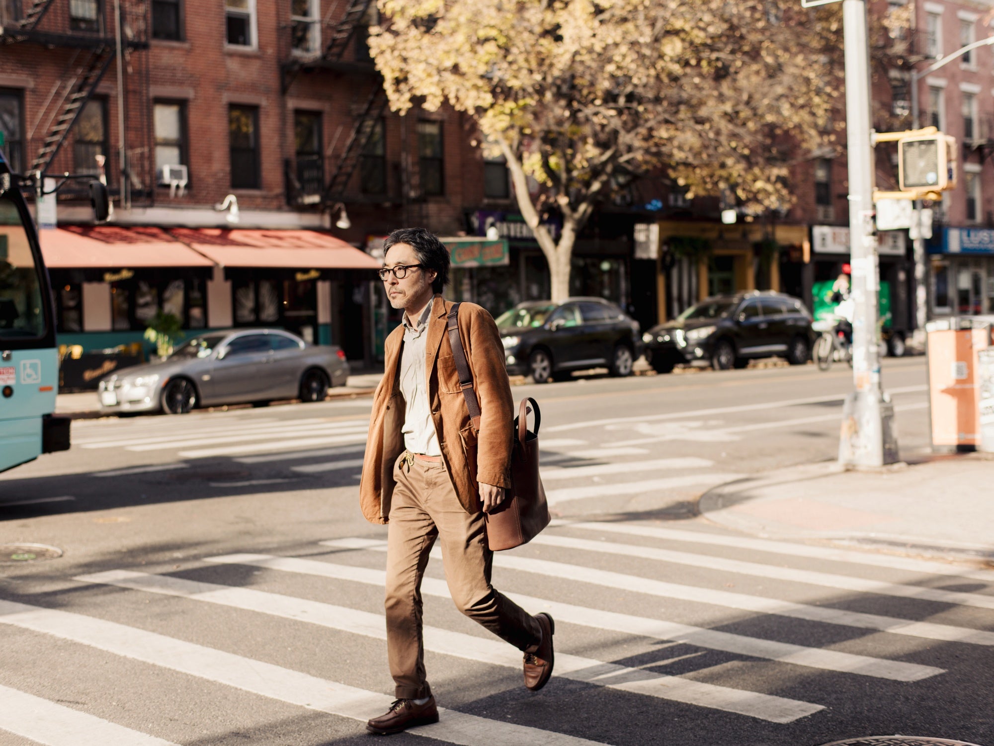 Kenzo Minami crosses a New York crosswalk while carrying Tsuchiya Kaban’s Tone Nume Box Tote in Medium Brown.