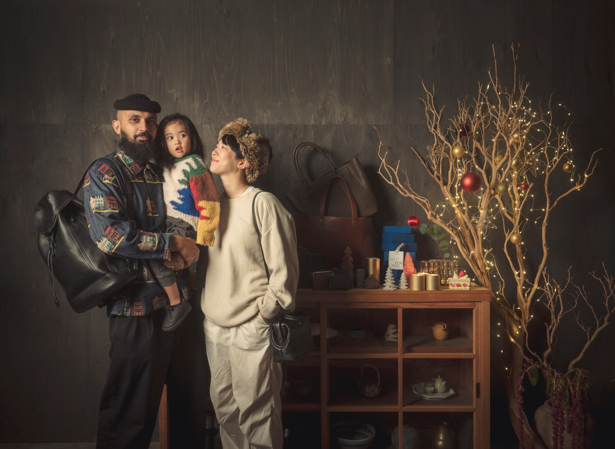 The Kanchan family of three poses for a photo in a cozy room. The father, Adhikari with a beard and a beret, holds a child in a colorful knit sweater. The mother, Asumi wearing a fuzzy winter hat, stands next to them. Behind them is a low wooden cabinet with various bags and gifts, and a small tree branch decorated with Christmas lights and ornaments.