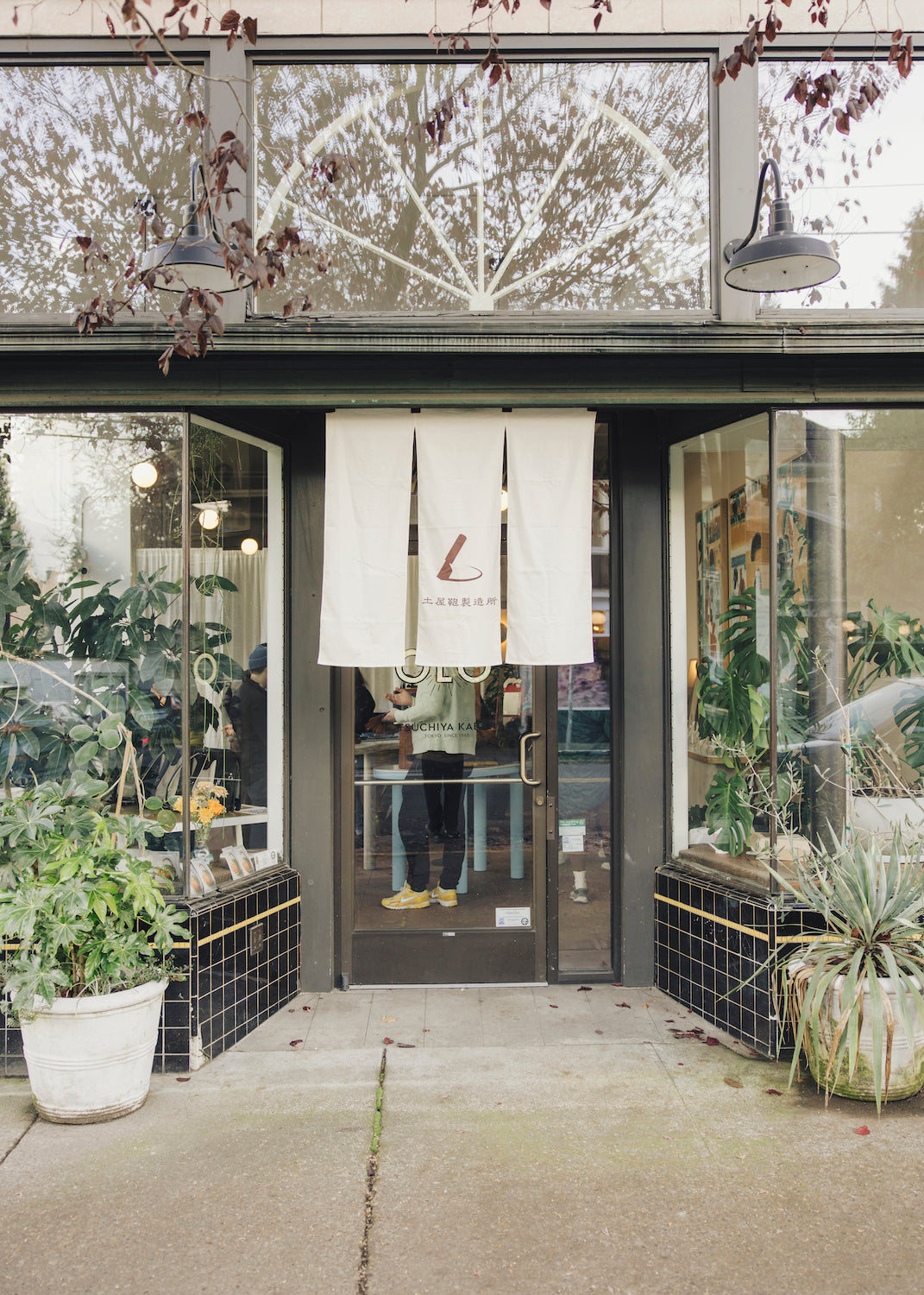 The entrance of a shop with a glass door and large windows, decorated with potted plants and featuring a white banner with a logo hanging above the door.