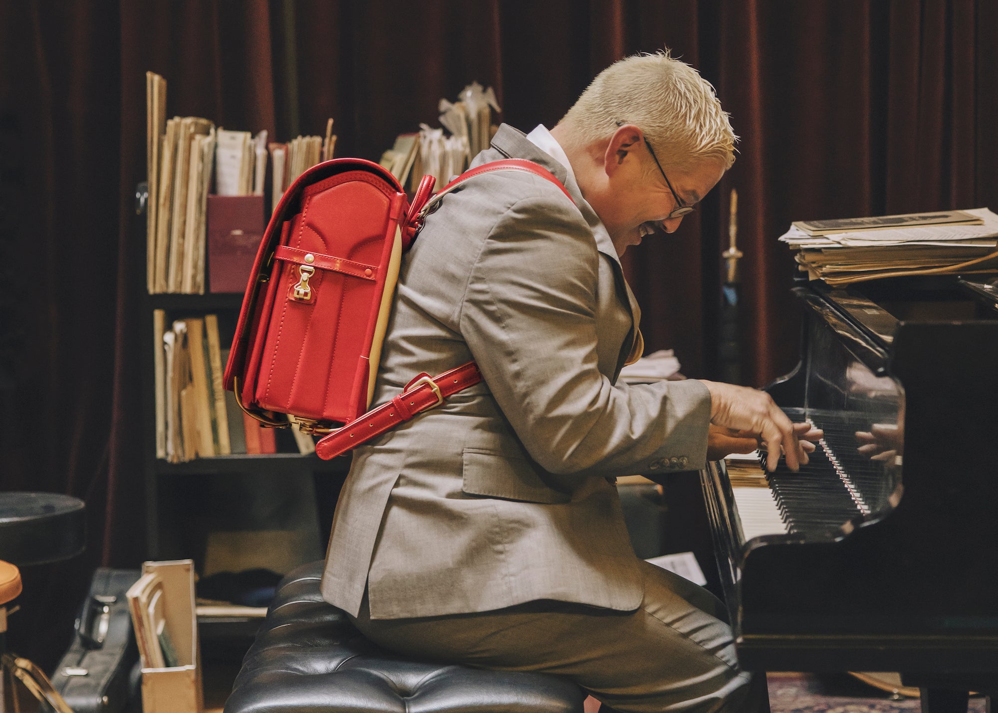 Thomas Lauderdale, wearing a suit and a red randoseru backpack, smiling while playing a piano.