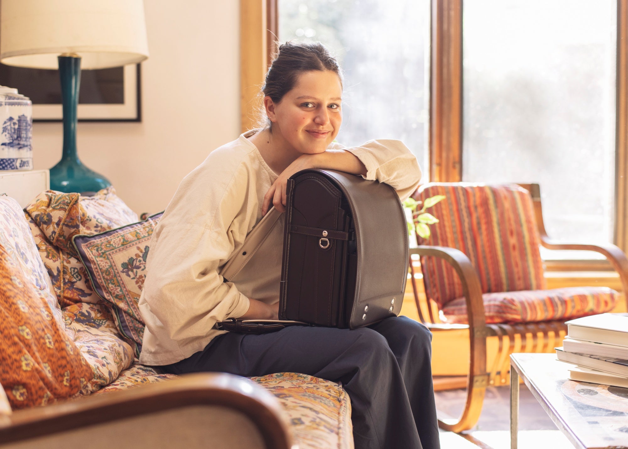 Megan Ladd with their hair tied back sits on a patterned couch, holding a Tsuchiya Kaban’s Randoseru in brown, structured bag on their lap. They are wearing a light-colored top and dark pants, and are smiling at the viewer. Behind them are a window with sunlight streaming in, a lamp, and a wooden armchair.