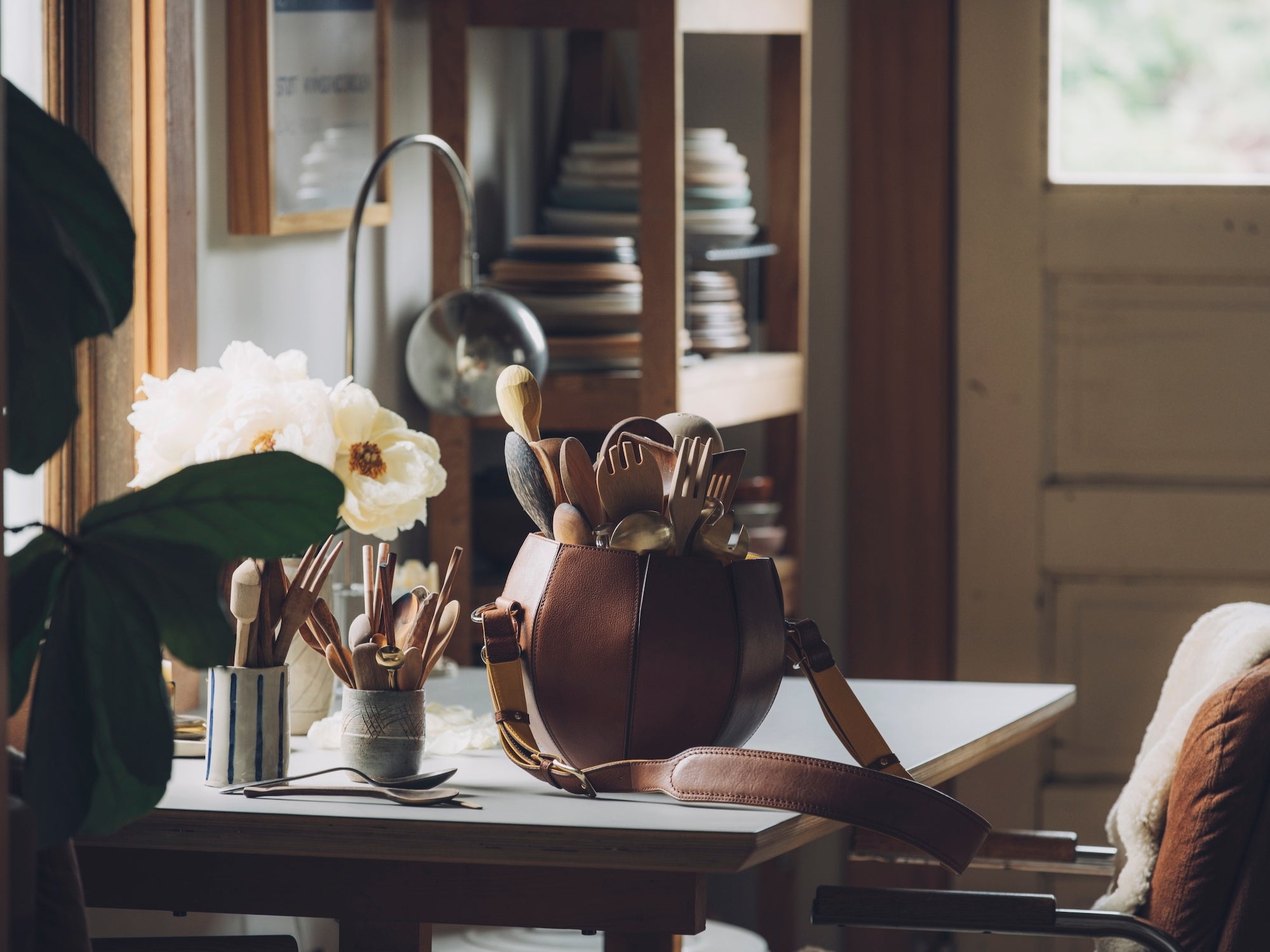 A round, brown leather bag with a handle is prominently displayed on a light-colored table. It is filled with an assortment of wooden utensils, including forks and spoons. To the left, a vase holds a bouquet of white and cream-colored flowers. Another container filled with wooden utensils sits next to the flowers. In the background, a warm-toned room is visible, with wooden shelves holding plates and other objects, and a window to the right. A plant leaf is visible in the foreground on the left.