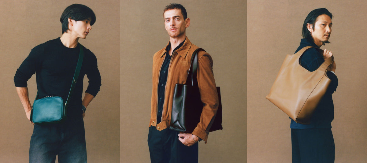 Three male models shown against a plain beige studio background, each wearing and carrying a leather shoulder bag in different colorways: a dark blue-green bag on the left, a brown-and-black bag in the center, and a large tan bag on the right.