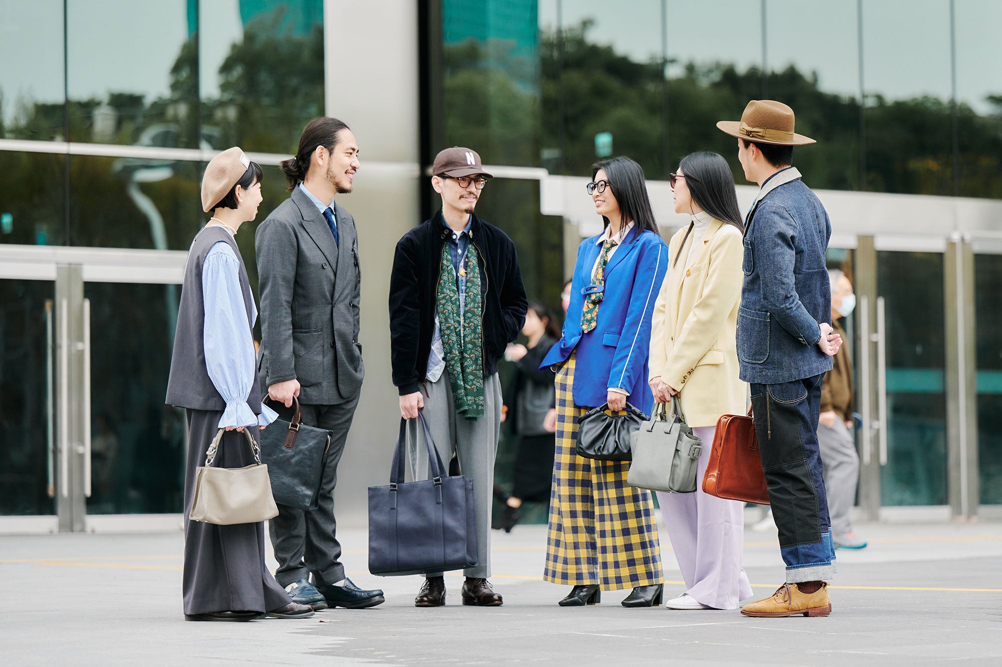 A group of six stylishly dressed Asian individuals are gathered outdoors on a paved area, possibly a plaza or a public space in front of a modern building with large glass windows reflecting the sky and surrounding environment.