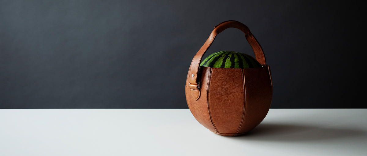 A unique brown leather handbag, shaped like a rounded bucket or a small watermelon, sits on a white surface against a dark gray background. A portion of a green, striped object, resembling the skin of a watermelon, is visible peeking out from the top of the bag. The handbag has a single, sturdy handle.