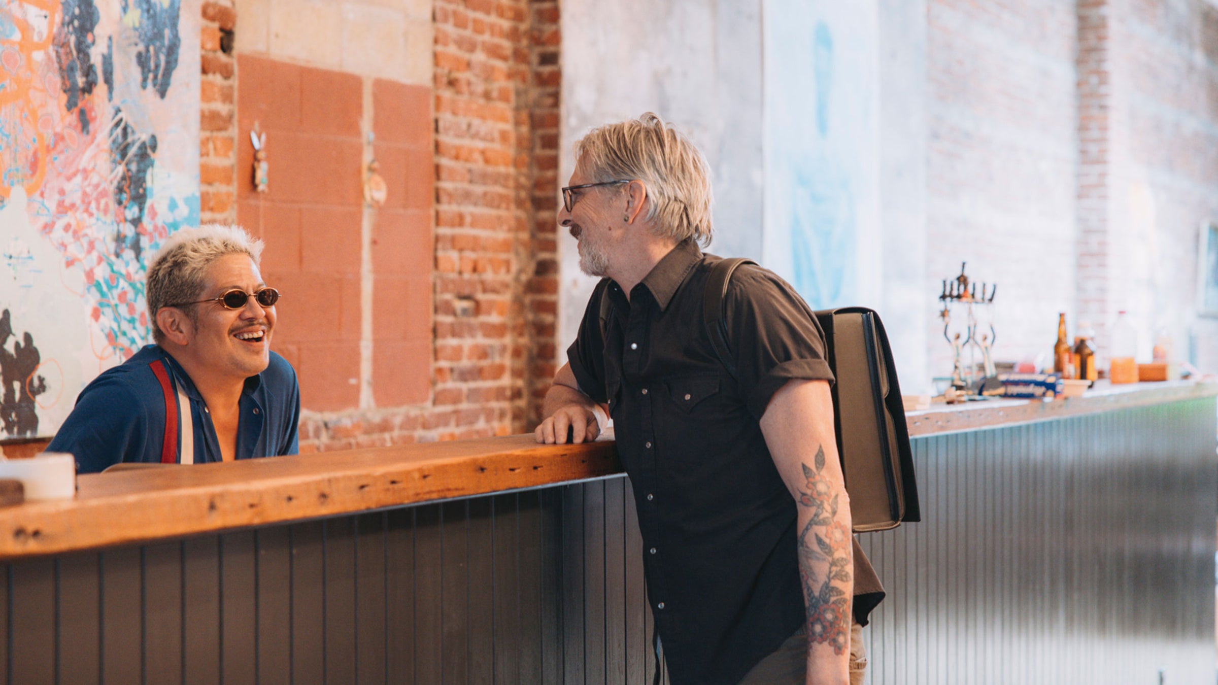 A vibrant indoor scene captures two men engaging in conversation across a wooden bar counter. On the left,  Thomas Lauderdale with light, curly hair and sunglasses, wearing a blue shirt with red suspenders, is smiling broadly and looking towards the other man. On the right, a man with graying hair and glasses, dressed in a dark short-sleeved shirt, is leaning on the counter and smiling back. He has a backpack slung over one shoulder and visible tattoos on his arm