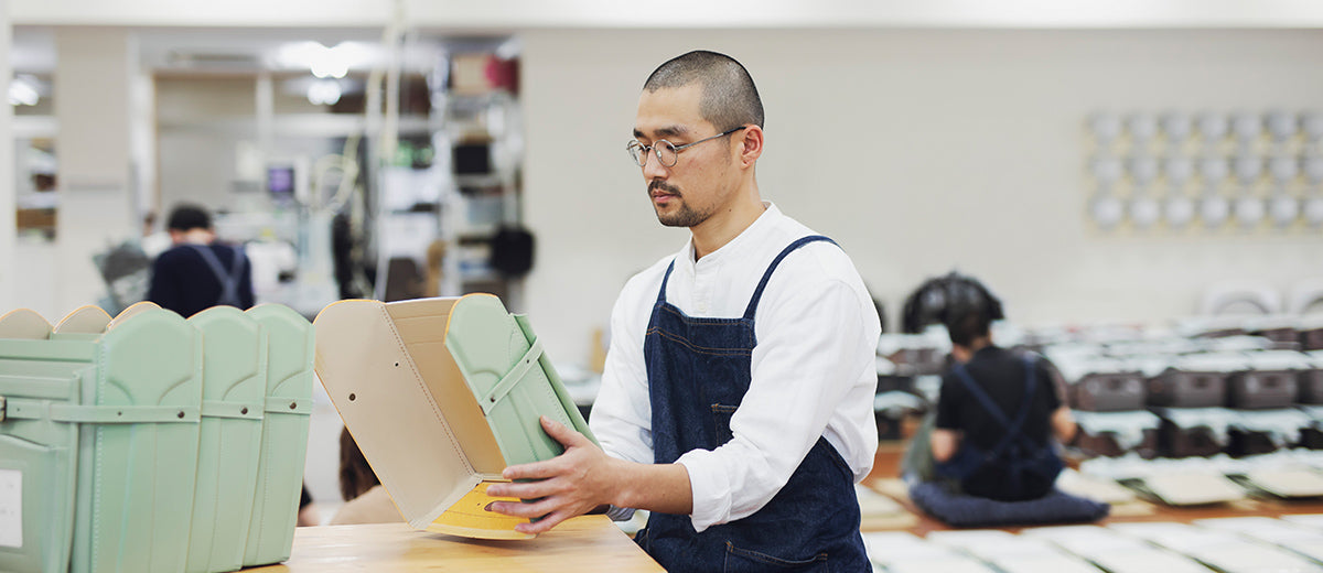 Suzuki san with short hair and glasses, wearing a white shirt and denim apron, is assembling a Tsuchiya Kaban’s Randoseru in a workshop.