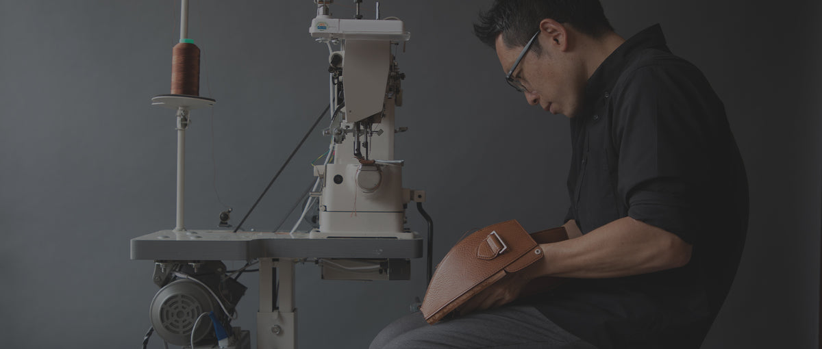 A person wearing glasses and a dark shirt is intently working on a brown leather item, possibly a bag or case, with both hands. They are seated in front of a white industrial sewing machine with a spool of brown thread visible to the left. The background is a plain, dark gray.