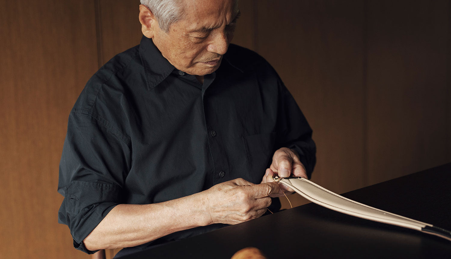 A focused shot shows Kunio Tsuchiya of Founder of Tsuchiya Kaban with gray hair, dressed in a dark, long-sleeved shirt, intently engaged in hand-stitching. He is holding a piece of light-colored material, possibly leather or heavy fabric, which has a dark edge, and threading a needle through it. His hands are visible, showing signs of age.