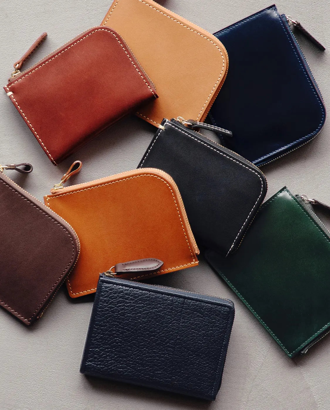 A group of eight small, rectangular leather wallets are arranged on a gray surface. Each wallet is a different color, including shades of brown, tan, navy, black, and green. They all have a similar zippered design with visible stitching and are spread out in a loose, overlapping pattern.